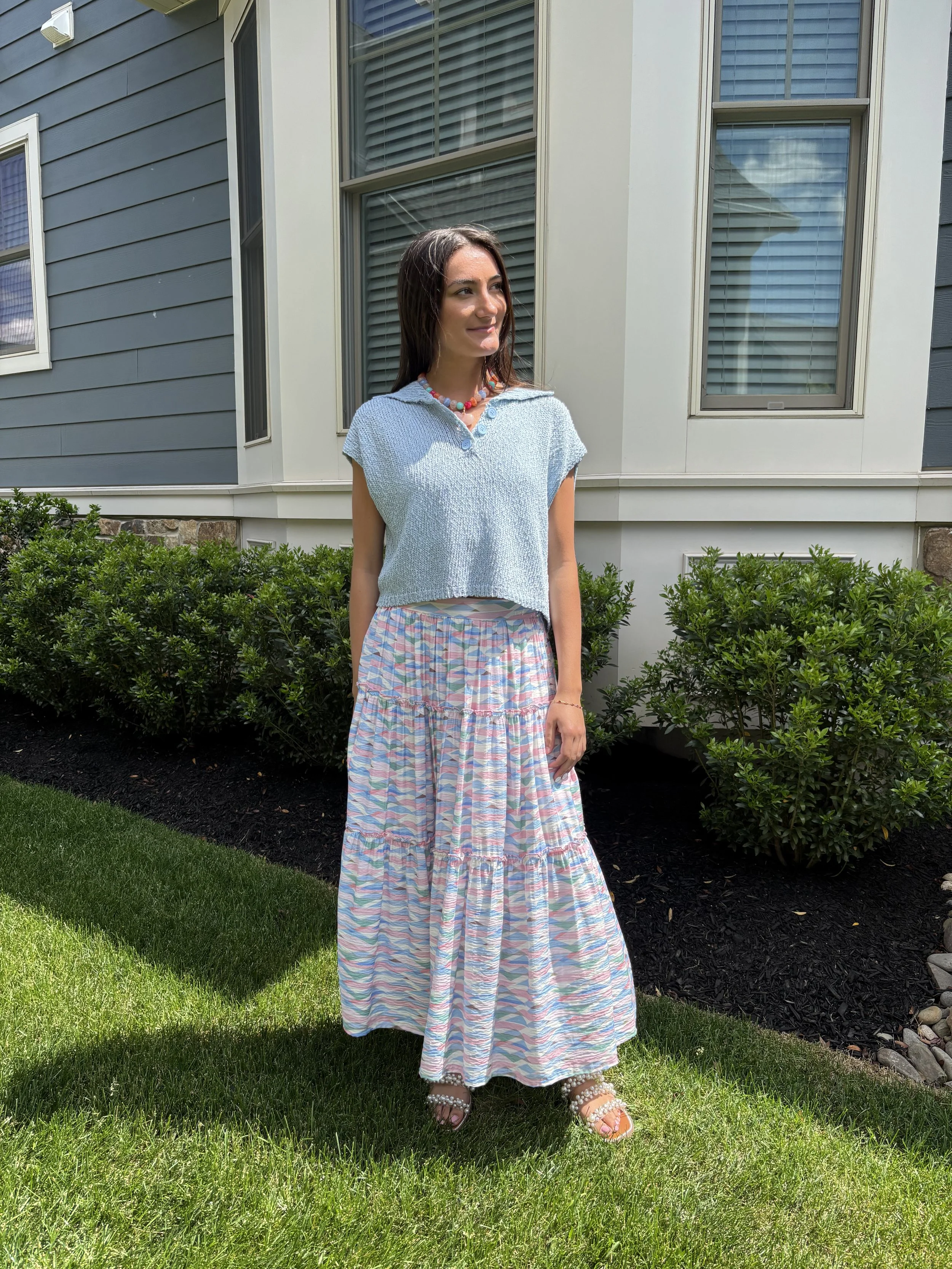 A woman standing outdoors in front of a house with large windows, wearing a light blue top with a collar, a colorful necklace, a long pastel-colored skirt, and sandals with beads, surrounded by green bushes and grass.