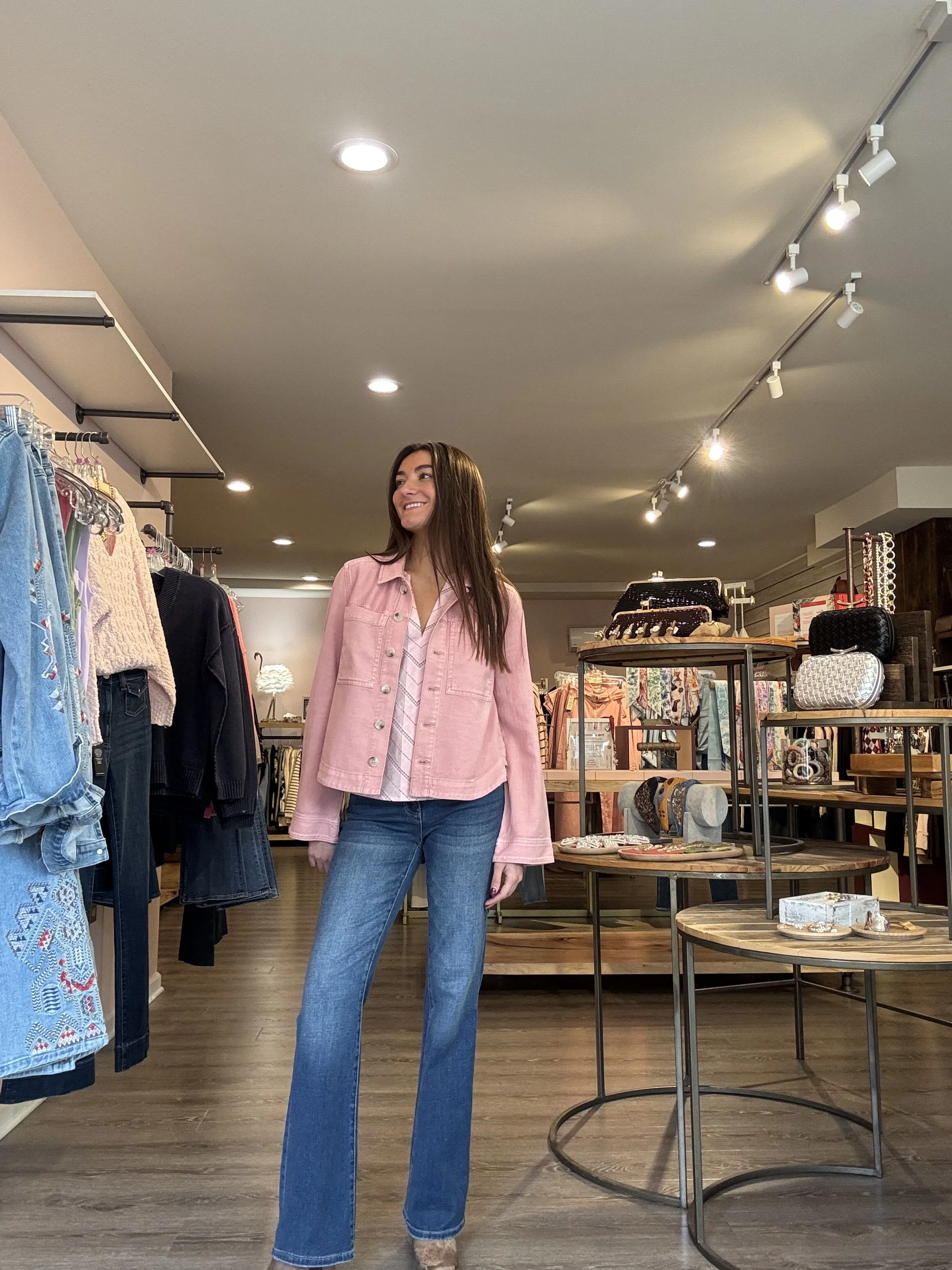 A young woman with long brown hair smiling while shopping in a boutique store, wearing a pink jacket and blue jeans.