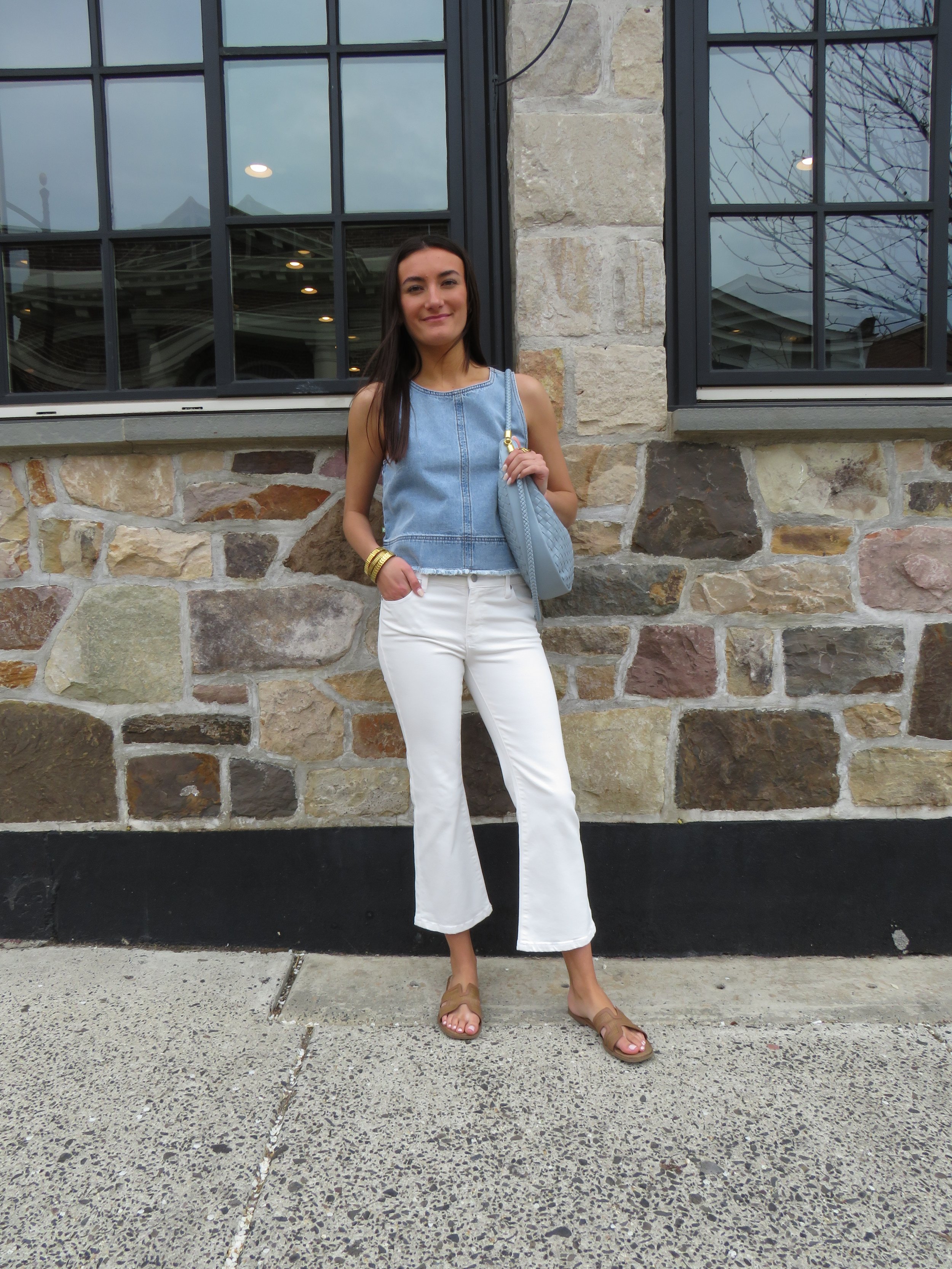 A woman standing in front of a stone and brick building with large black framed windows, wearing a sleeveless denim top, white pants, tan sandals, and holding a light blue quilted purse with a shoulder strap.