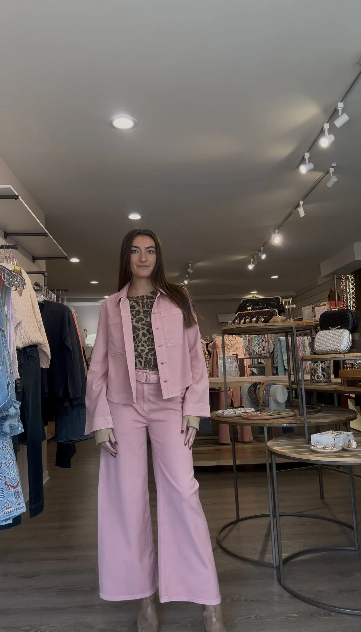 A young woman with long brown hair wearing a pink denim jacket and matching wide-leg pants, standing inside a clothing store.