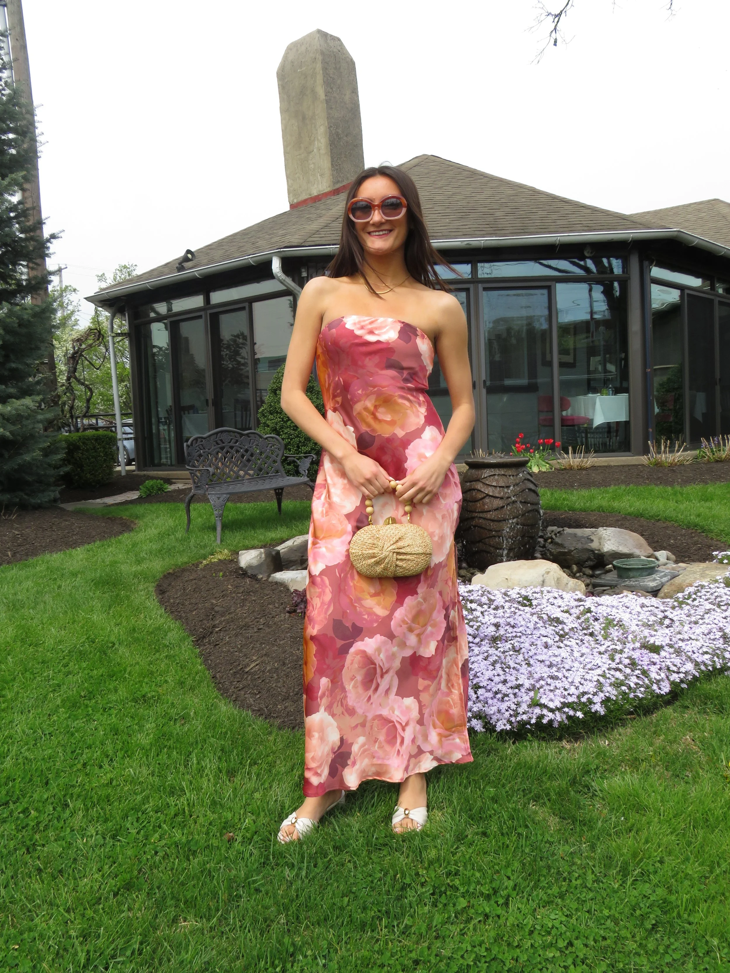 A woman with long dark hair wearing sunglasses and a strapless floral dress holding a small handbag, standing on a green lawn in front of a house with large windows and a garden, during daytime.