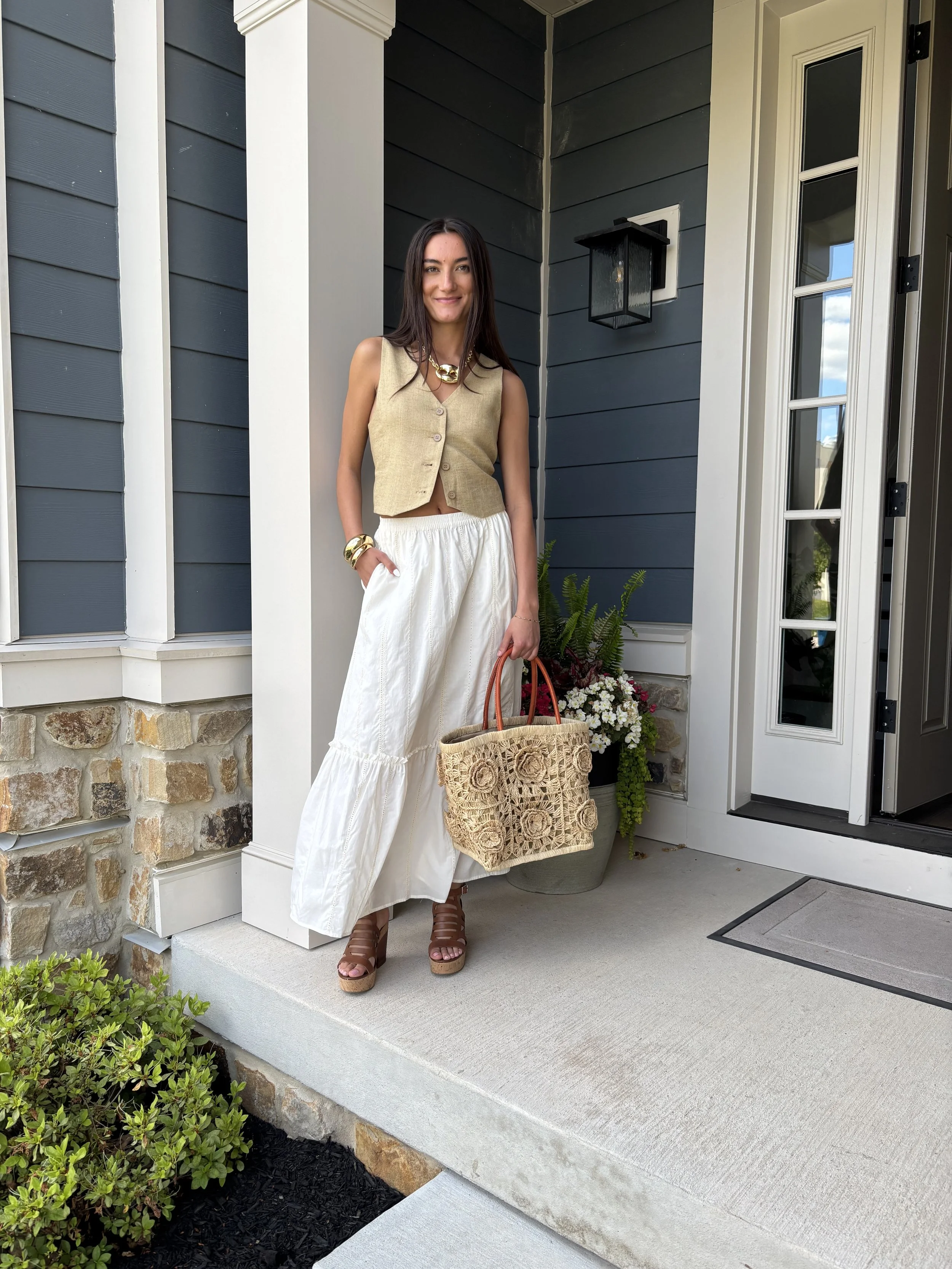 A young woman with long dark hair standing on a porch near a front door, holding a woven bag, dressed in a beige top and white skirt, wearing high heels and jewelry, with a potted plant and flowers nearby.