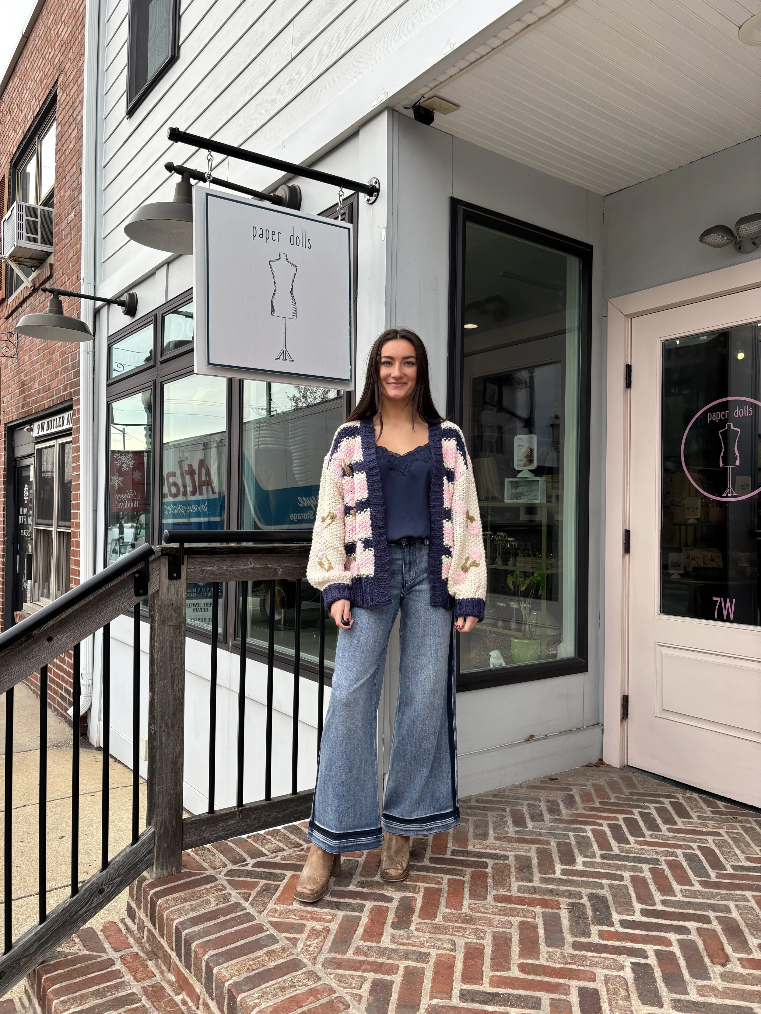 Young woman standing outside a store called paper dolls, wearing a navy blouse, wide-leg jeans, a colorful knitted cardigan, and beige boots.
