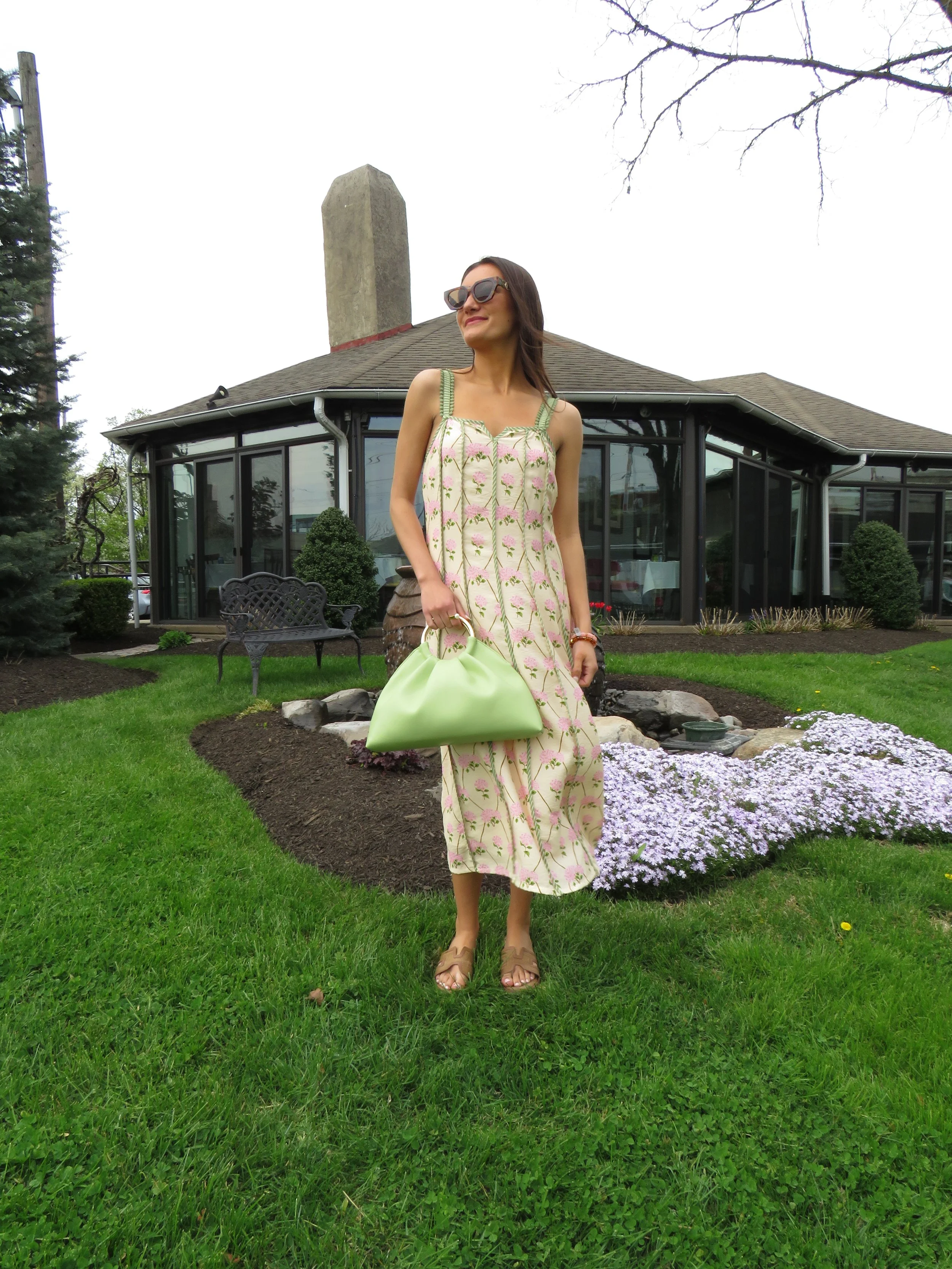 A woman in a flowery dress and sunglasses standing on a well-manicured lawn with a garden behind her, a house with large windows, and a leafless tree in the background.