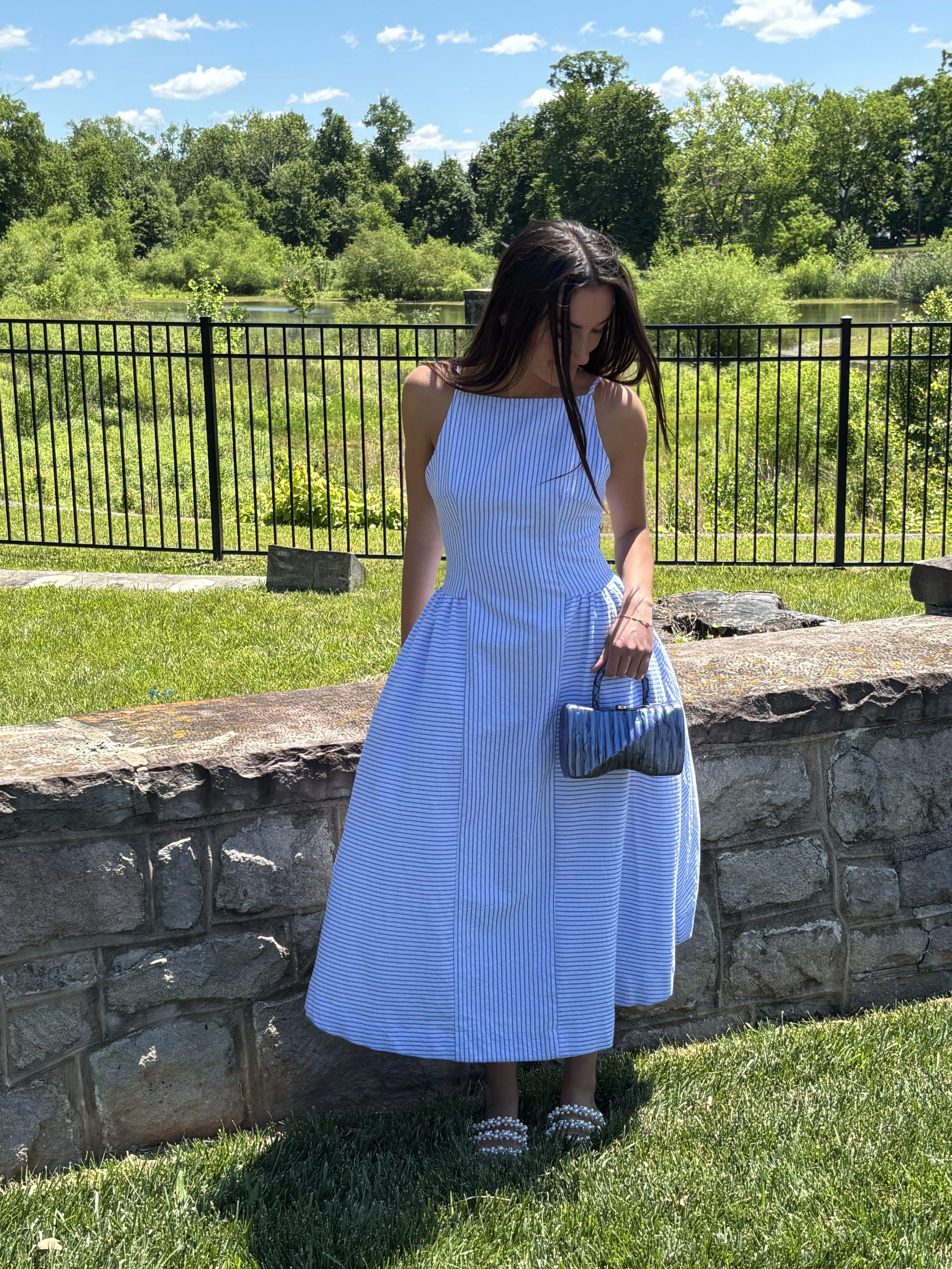A woman wearing a blue and white striped dress standing by a stone wall near a grassy area with a lake and trees in the background on a sunny day.