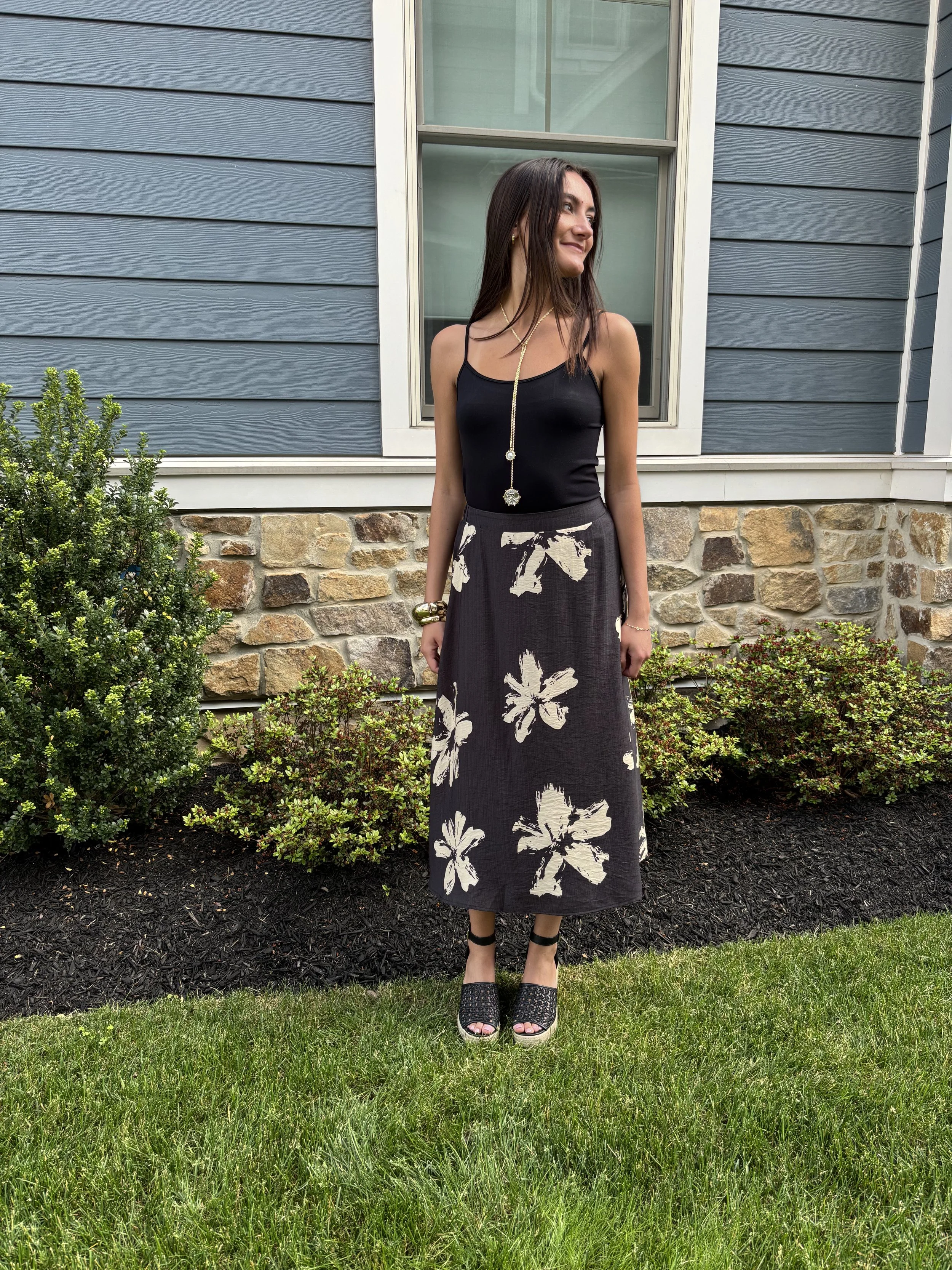 A woman in a black tank top and floral printed skirt standing outdoors in front of a house with blue siding and a stone foundation. She is smiling and wearing jewelry and black platform shoes.
