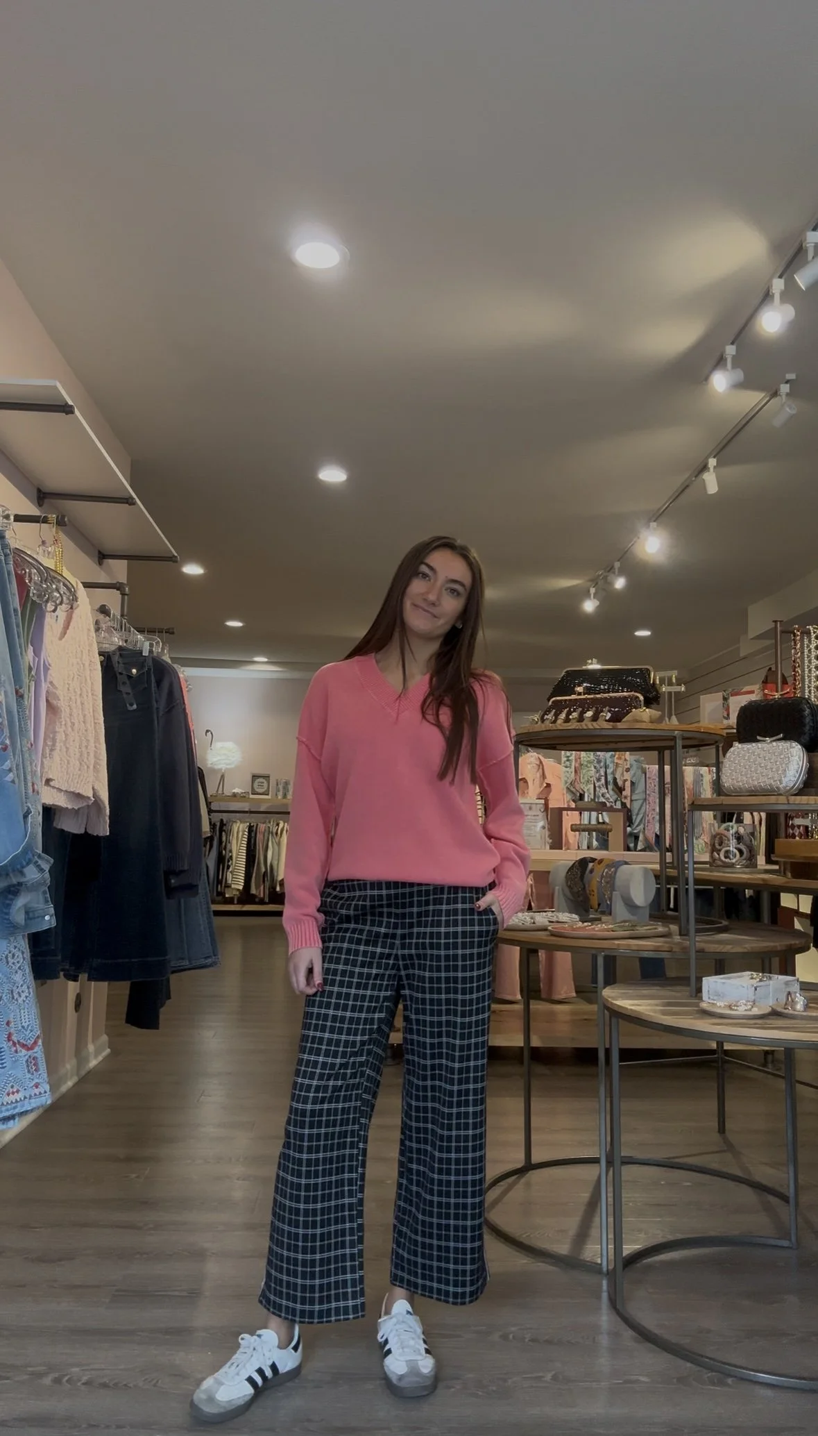 A young woman with long brown hair standing in a clothing store, wearing a pink sweater, checkered wide-leg pants, and white sneakers with black stripes, surrounded by racks and shelves of clothing and accessories.