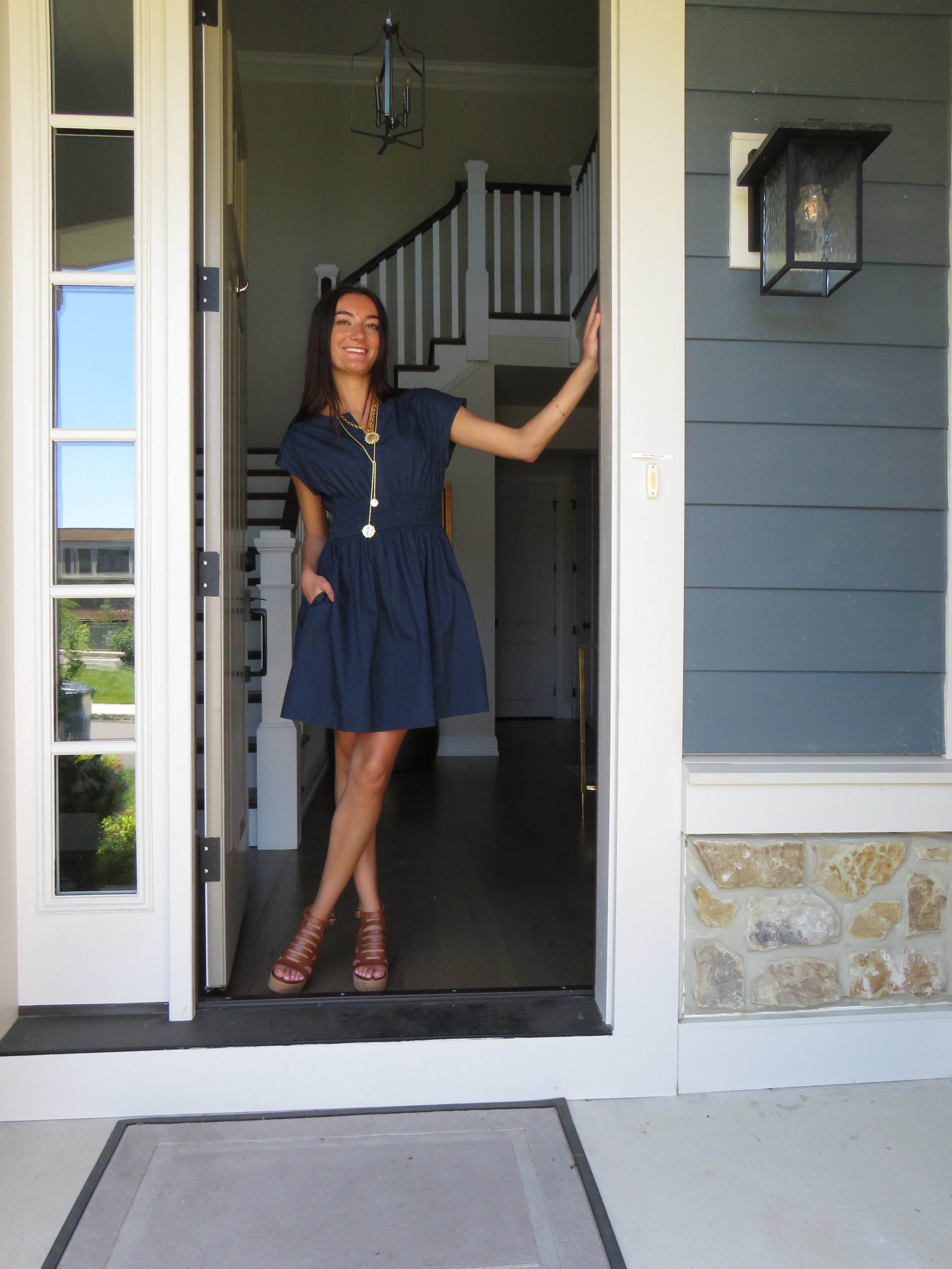 A young woman in a blue dress and brown high heels standing at the entrance of her house, touching the doorframe with her right hand, smiling.