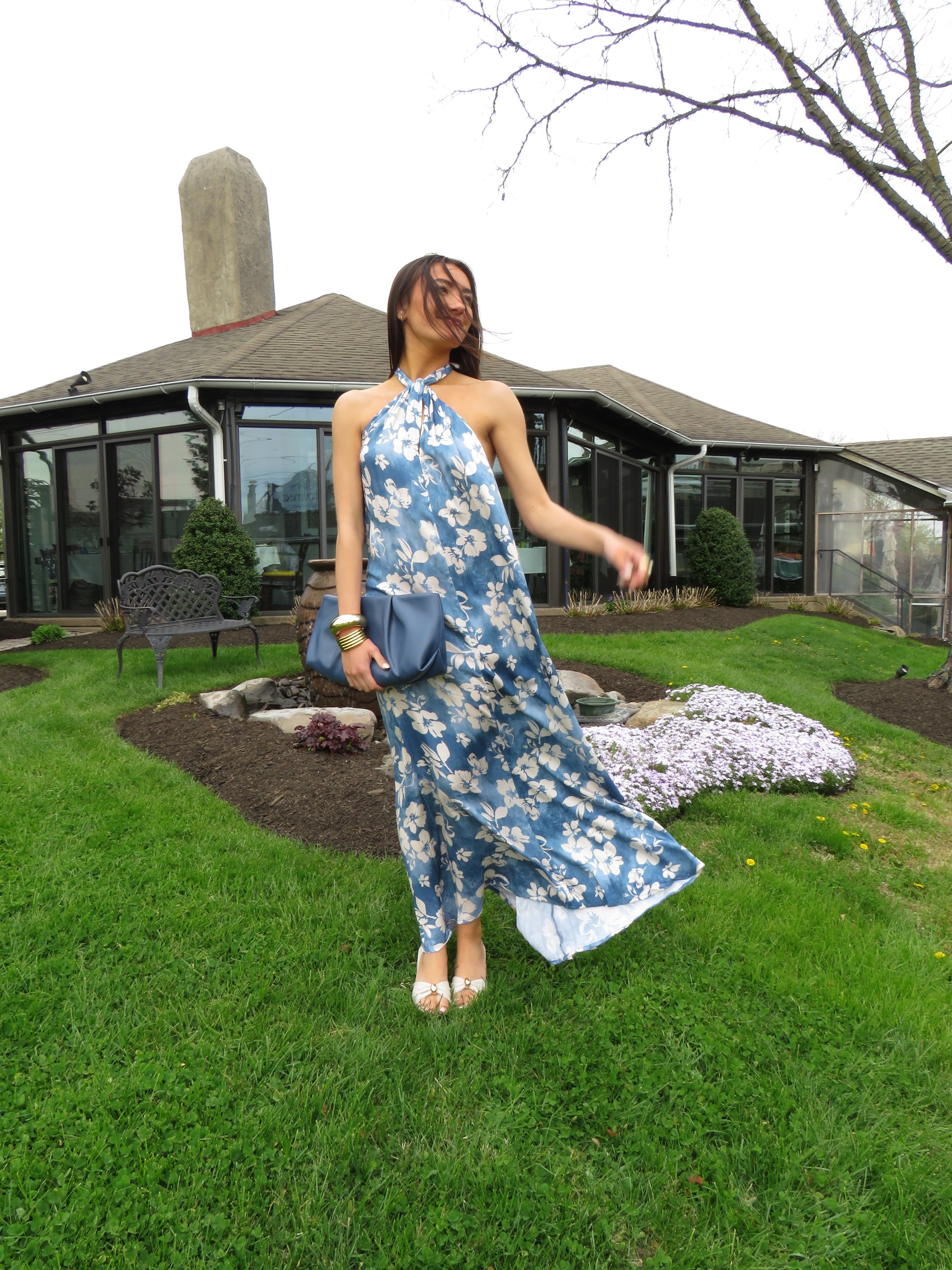 A woman in a blue and white floral dress standing in a garden with green grass, holding a blue purse, and smiling with her hair blowing in the wind, in front of a house with large windows and trimmed bushes.