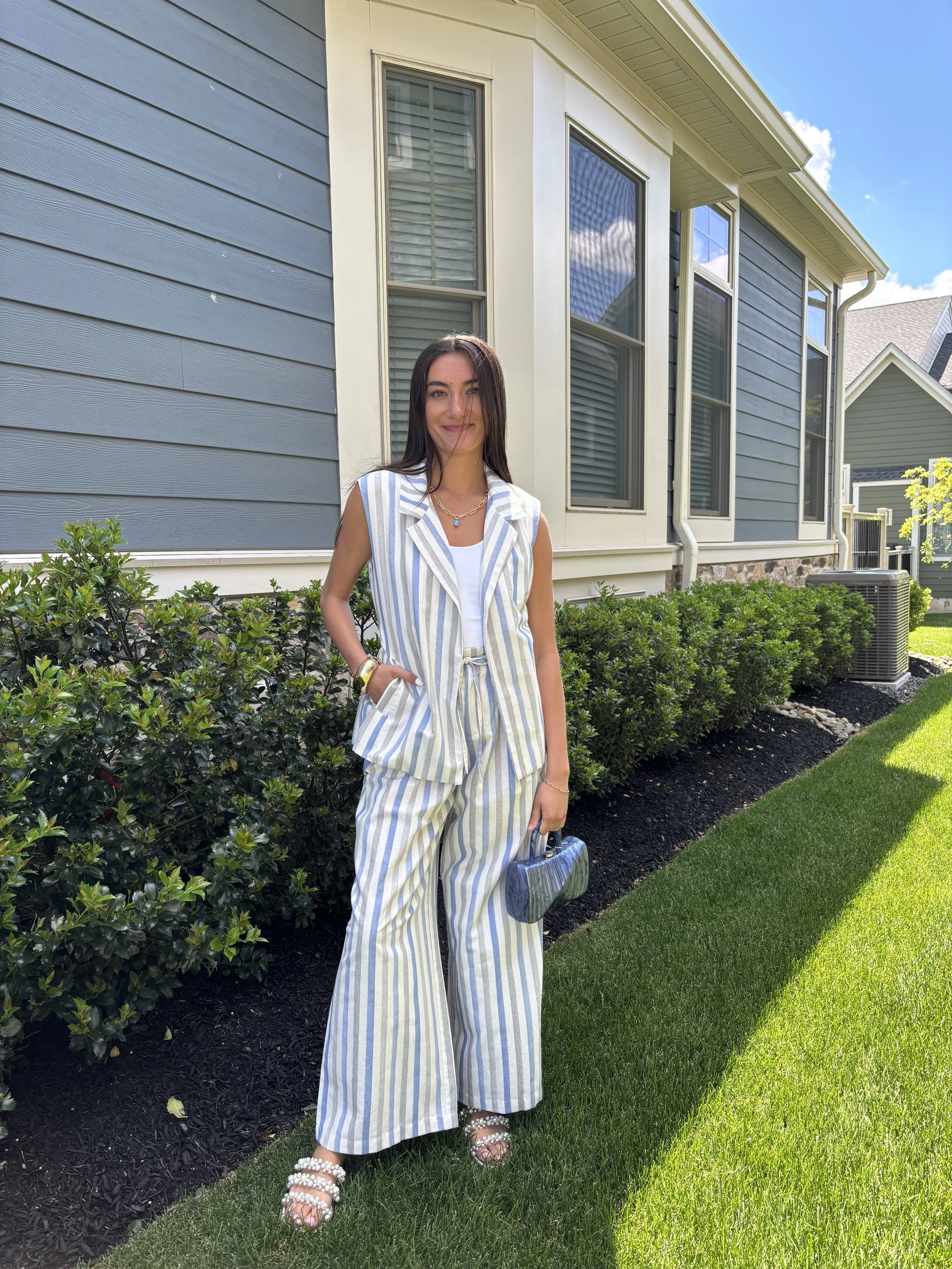 A young woman in wide-leg striped pants and a matching sleeveless blazer, holding a small purse, standing on a lush green lawn in front of a modern house with large windows and bushes, on a sunny day.