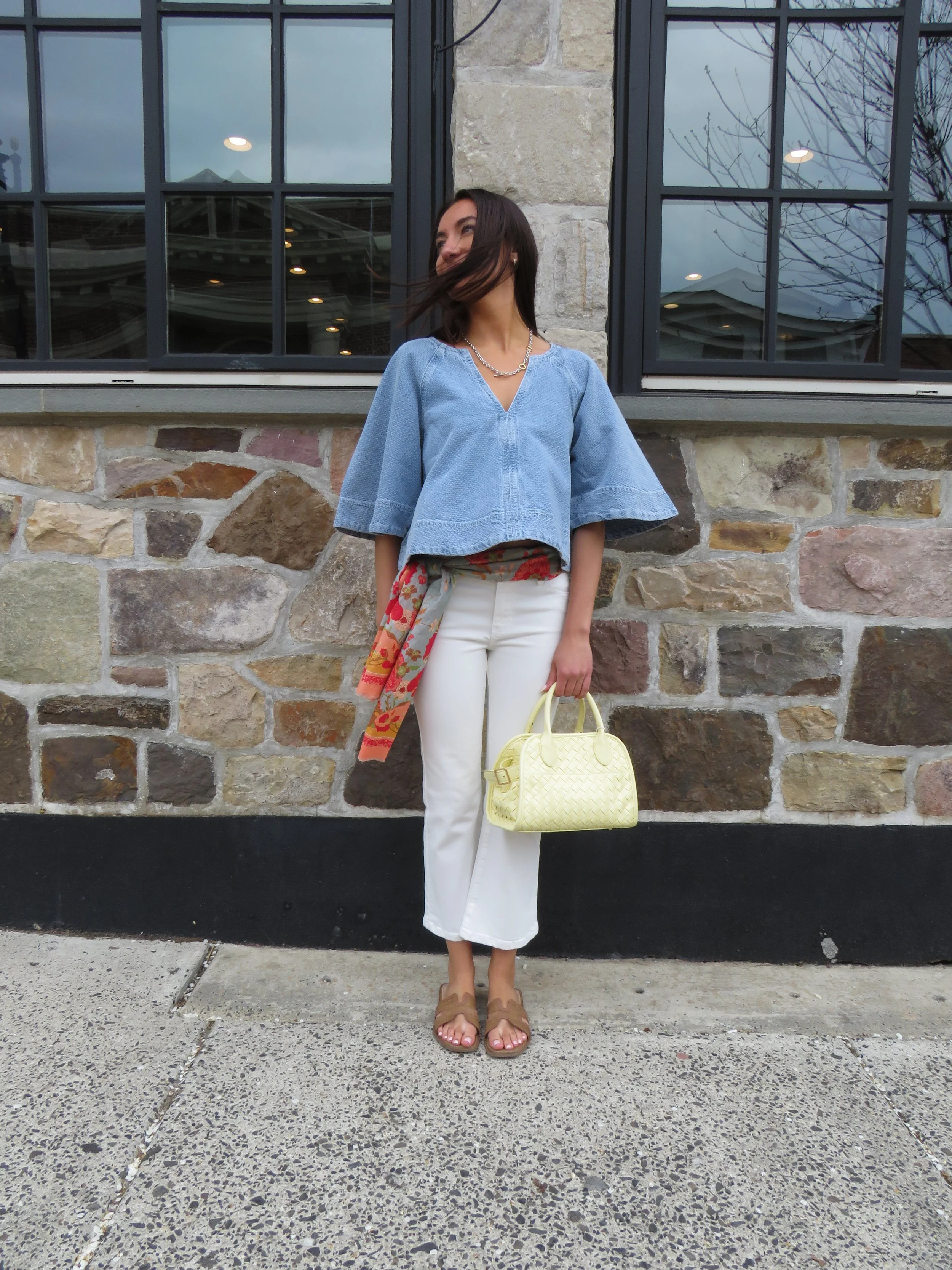 A woman standing against a stone wall with a large window behind her, wearing a blue blouse, white pants, brown sandals, and holding a yellow handbag.