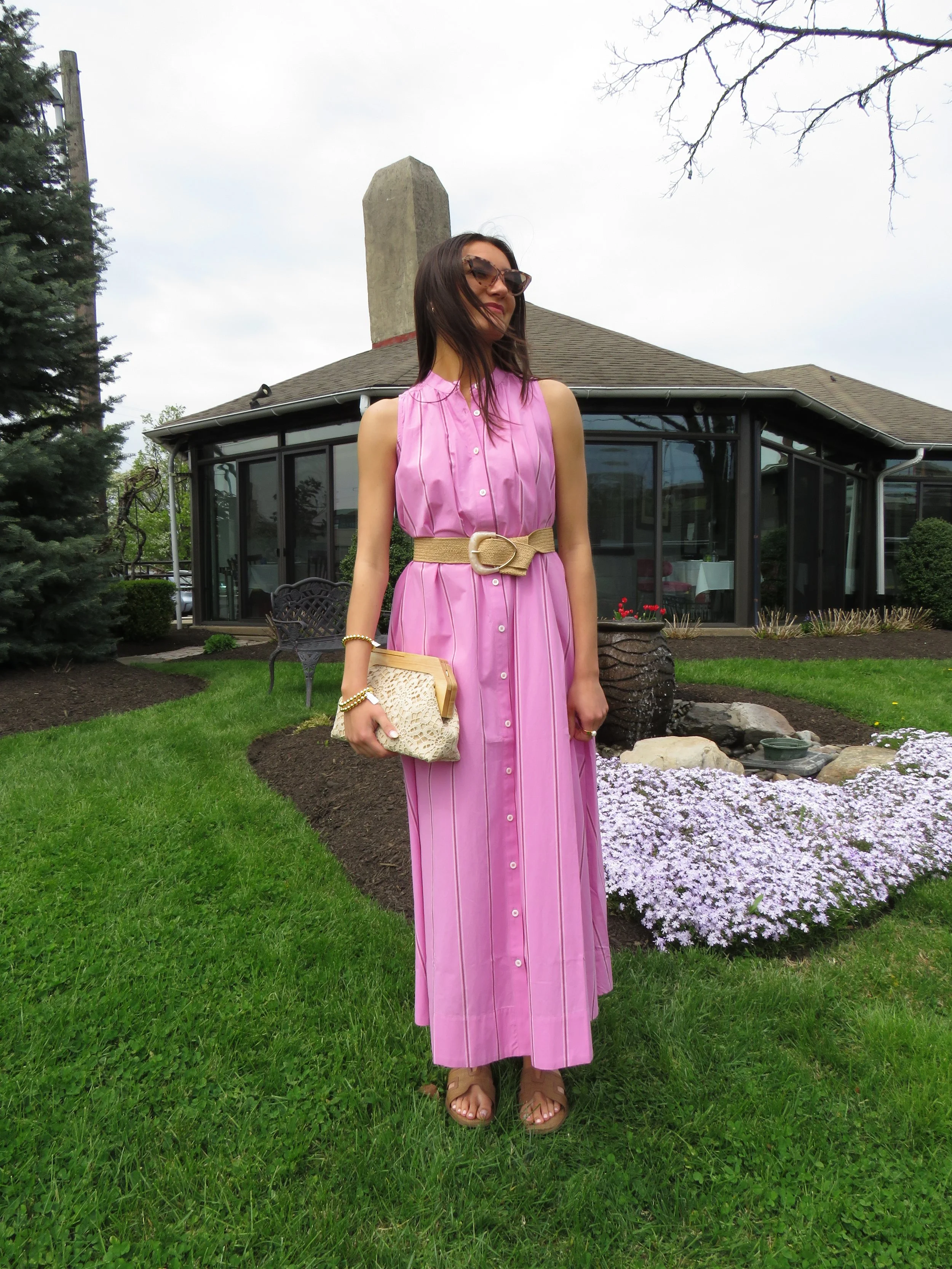 Woman in a pink dress and sunglasses standing in a garden with purple flowers and a house in the background.