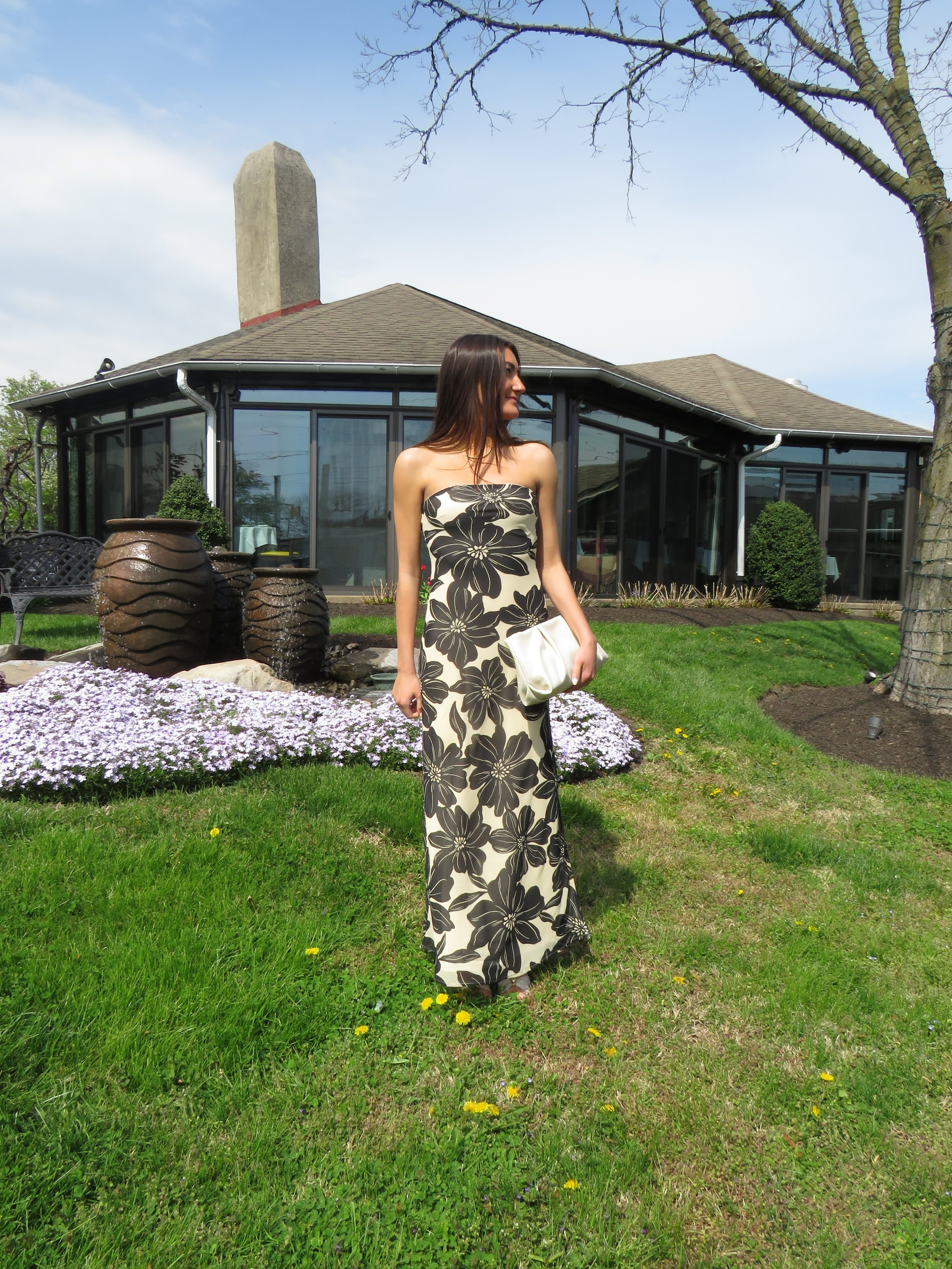 A woman in a strapless black and white floral dress standing outside in a garden with green grass, yellow flowers, a tree, and large clay pots, with a house featuring large windows and a chimney in the background.