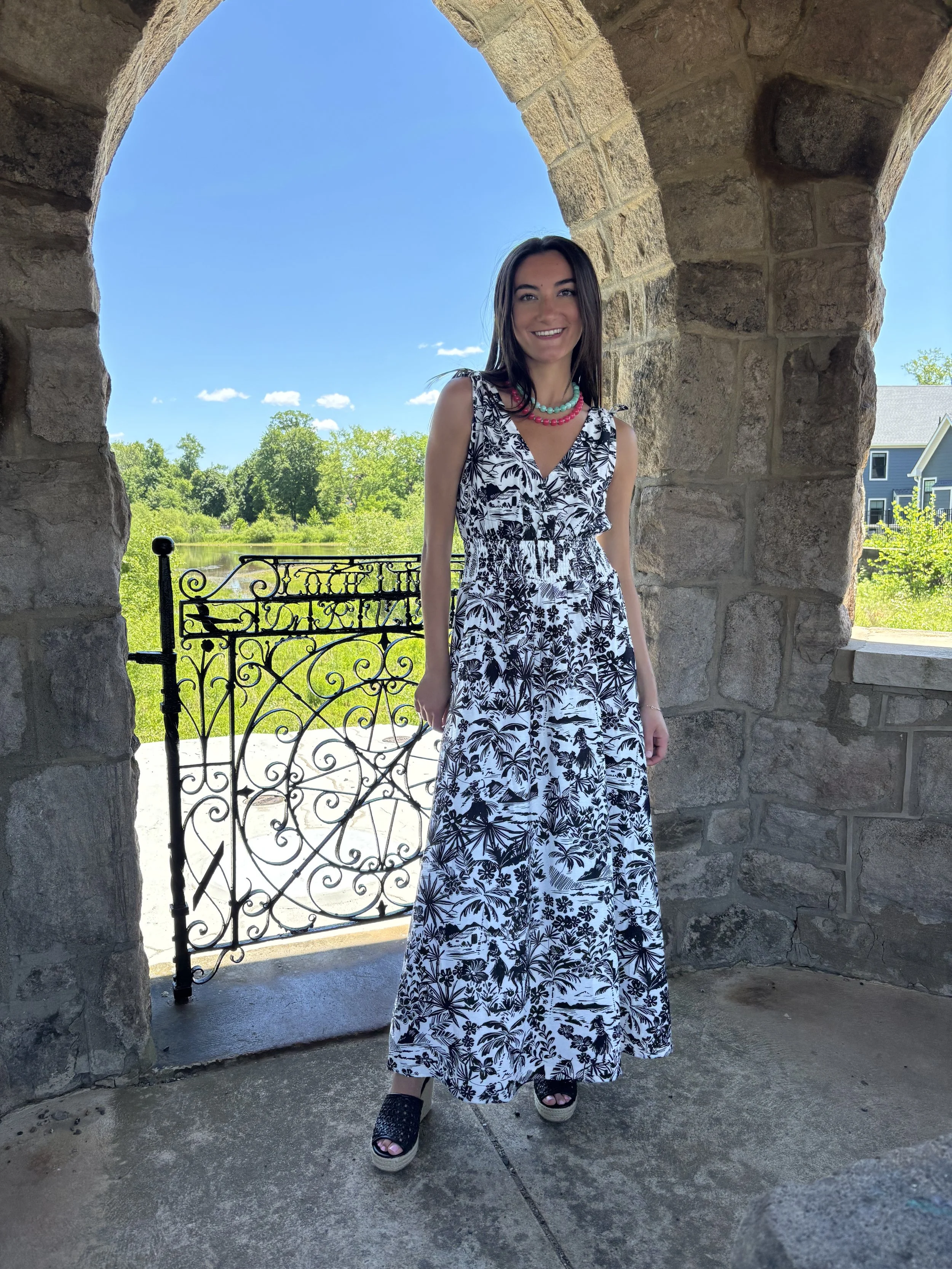 A woman wearing a black and white floral maxi dress and platform sandals standing in front of a stone archway, with lush greenery and a blue sky in the background.