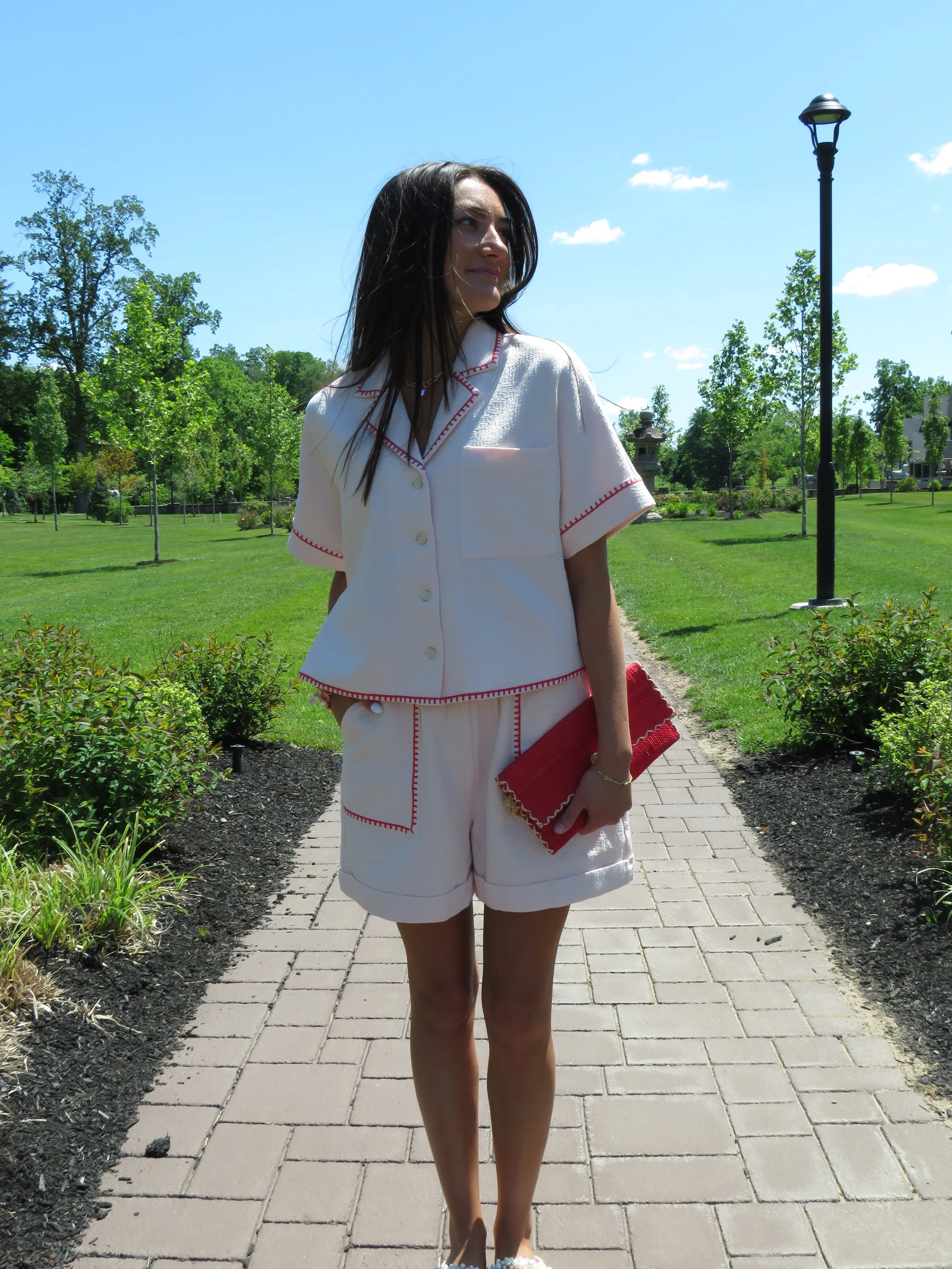 A woman with dark hair wearing a white matching pajama set with red stitching, holding a red clutch, standing on a brick pathway in a garden with green trees and a lamppost, under a bright blue sky with a few clouds.