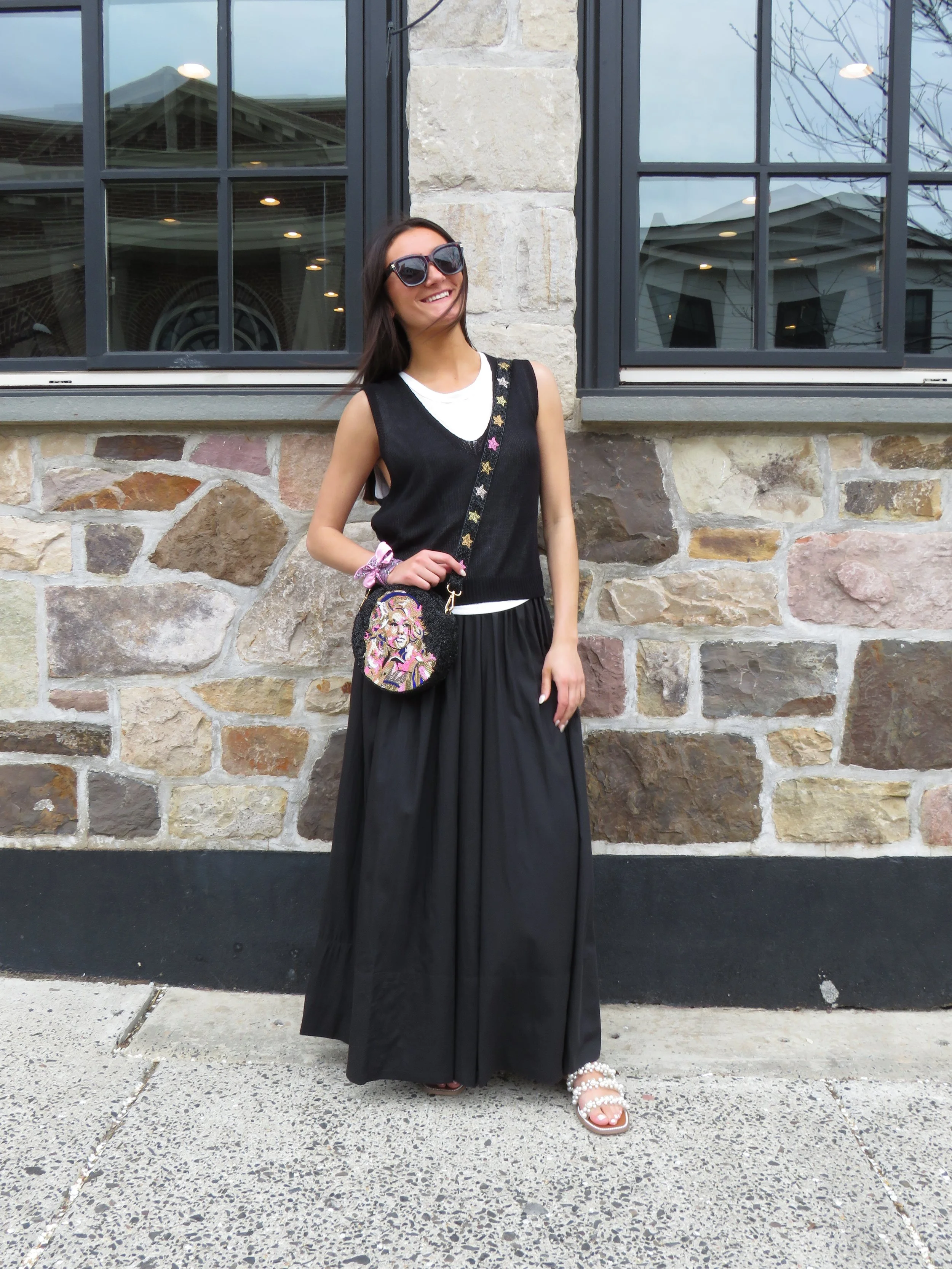A woman standing in front of a stone building with large windows, wearing sunglasses, a black sleeveless sweater with colorful star details, a long black skirt, and pearl-embellished sandals.
