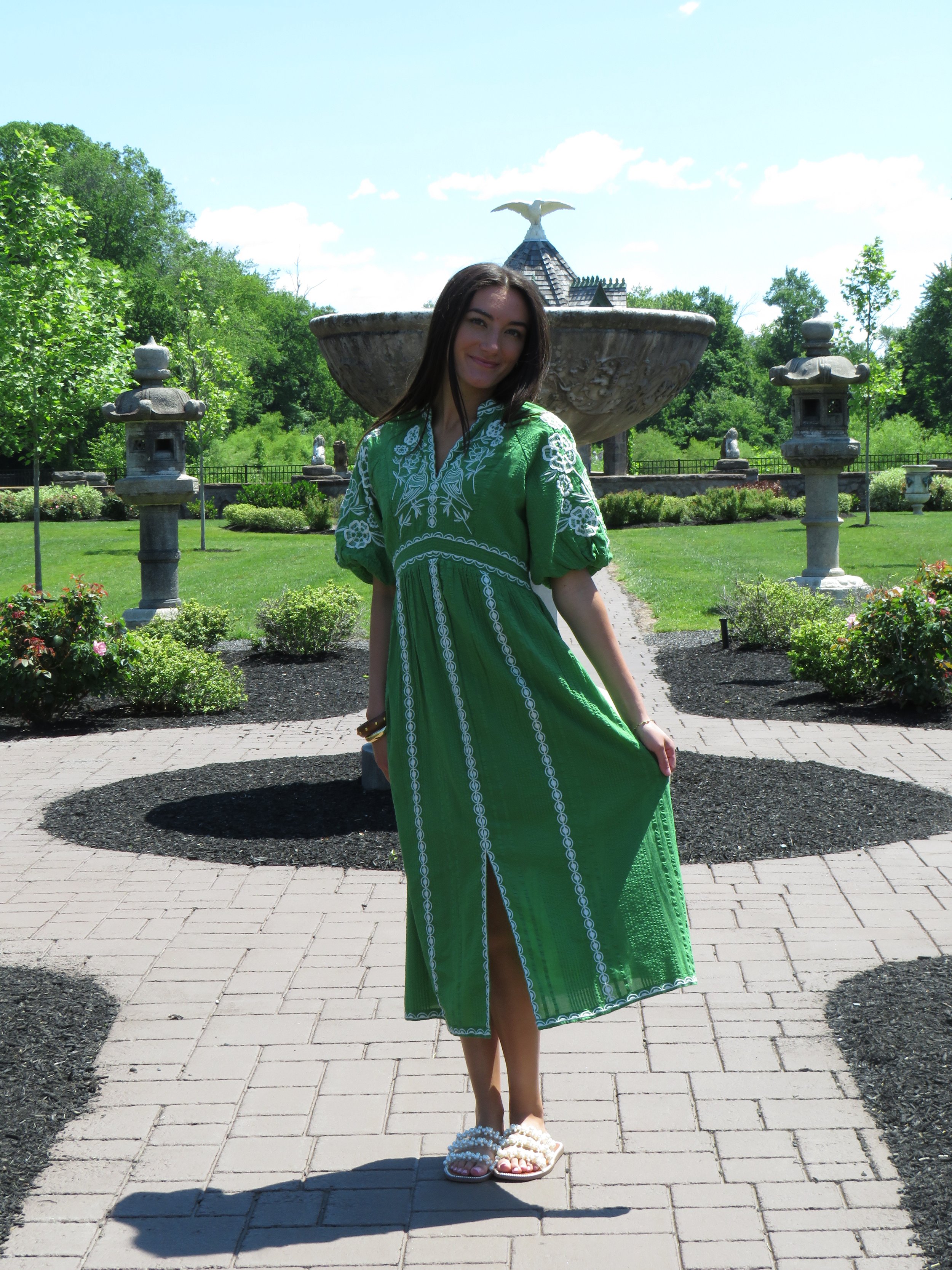 Young woman in a green dress standing on a brick path in front of a large fountain with an eagle sculpture on top, surrounded by greenery and traditional stone lanterns.