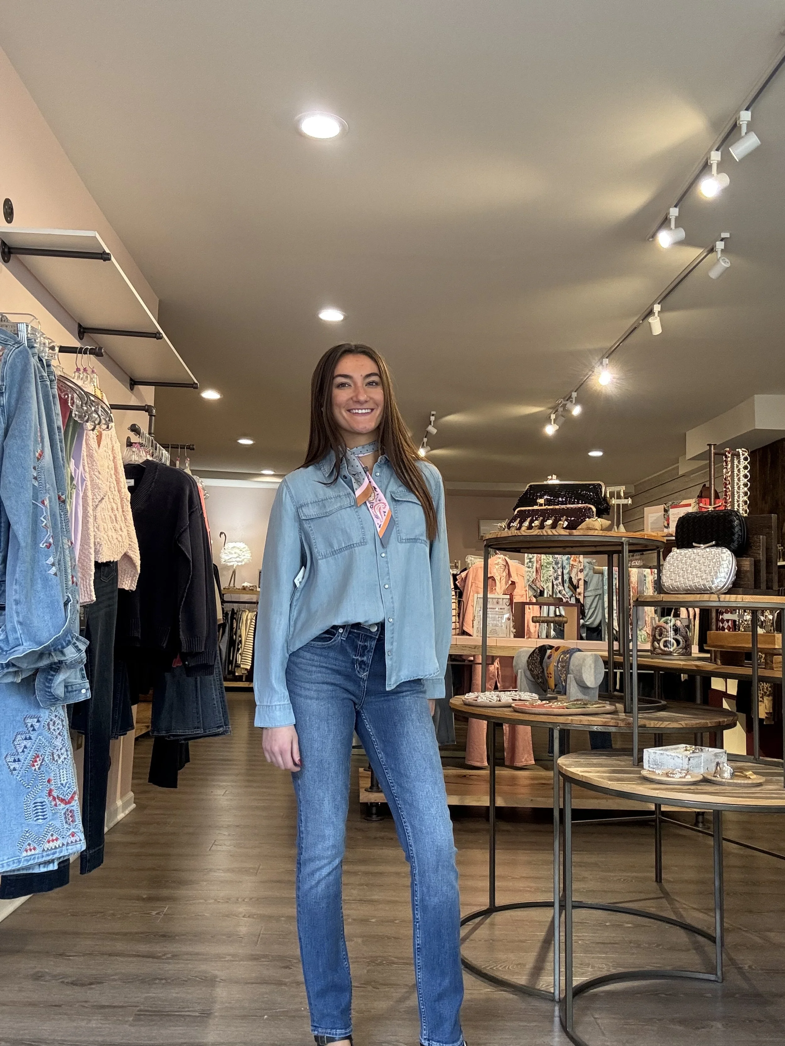 A young woman standing inside a clothing store, smiling at the camera. She is wearing a denim jacket, jeans, and a patterned bandana around her neck. The store has various racks of clothing and accessories displayed on tables and shelves.