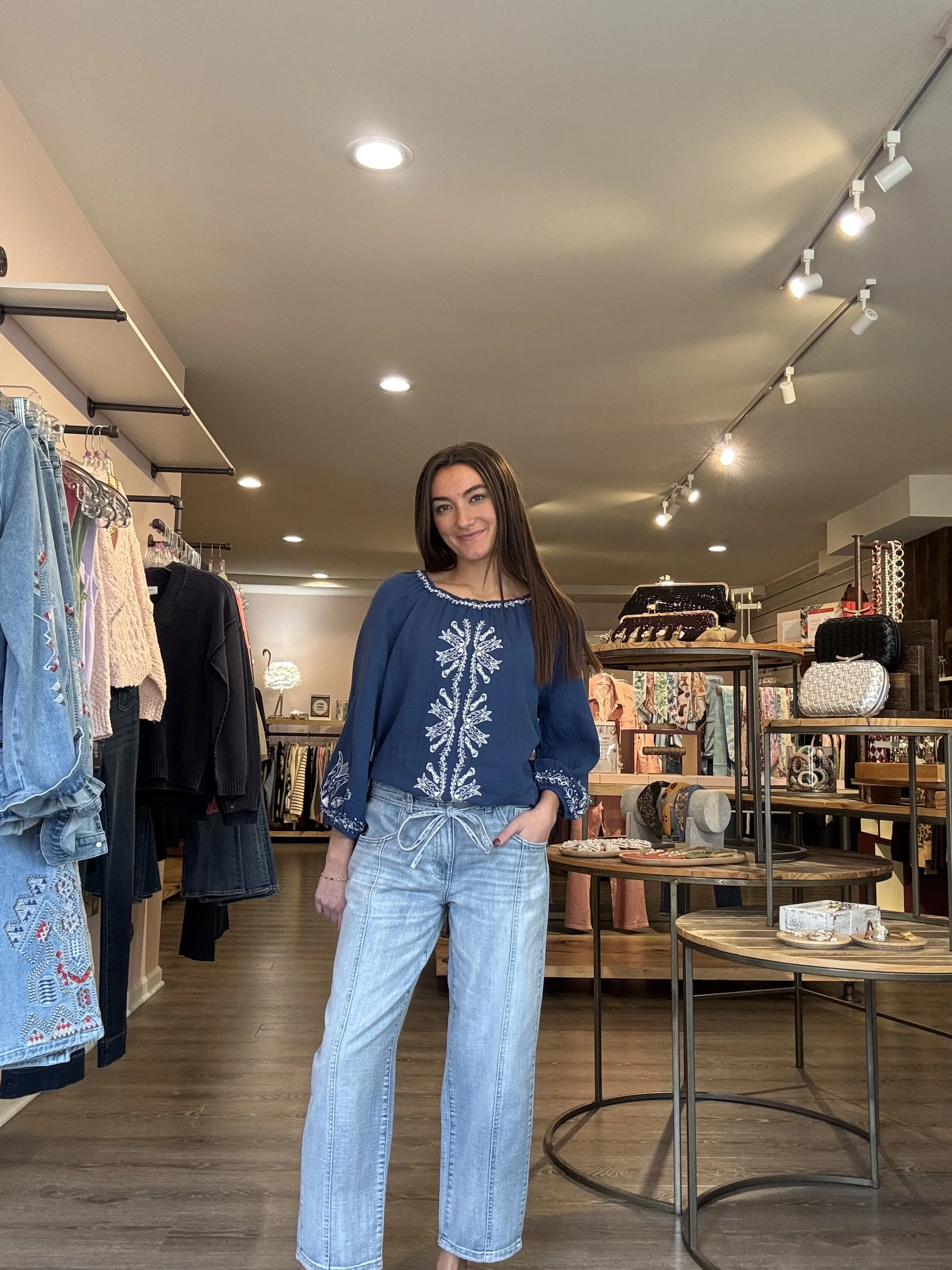 A young woman standing in a clothing store, smiling at the camera, wearing a blue embroidered blouse and light blue jeans.