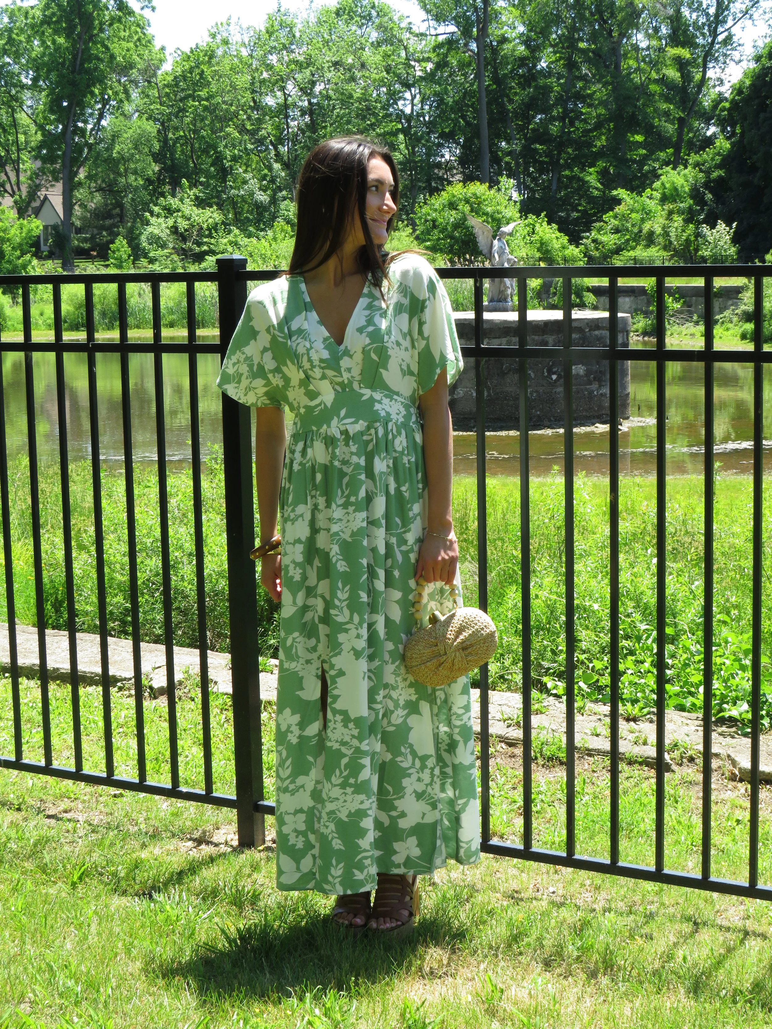 A woman with dark hair wearing a green and white floral dress holding a straw hat, standing outdoors by a black metal fence near a pond with a statue and lush green trees in the background.