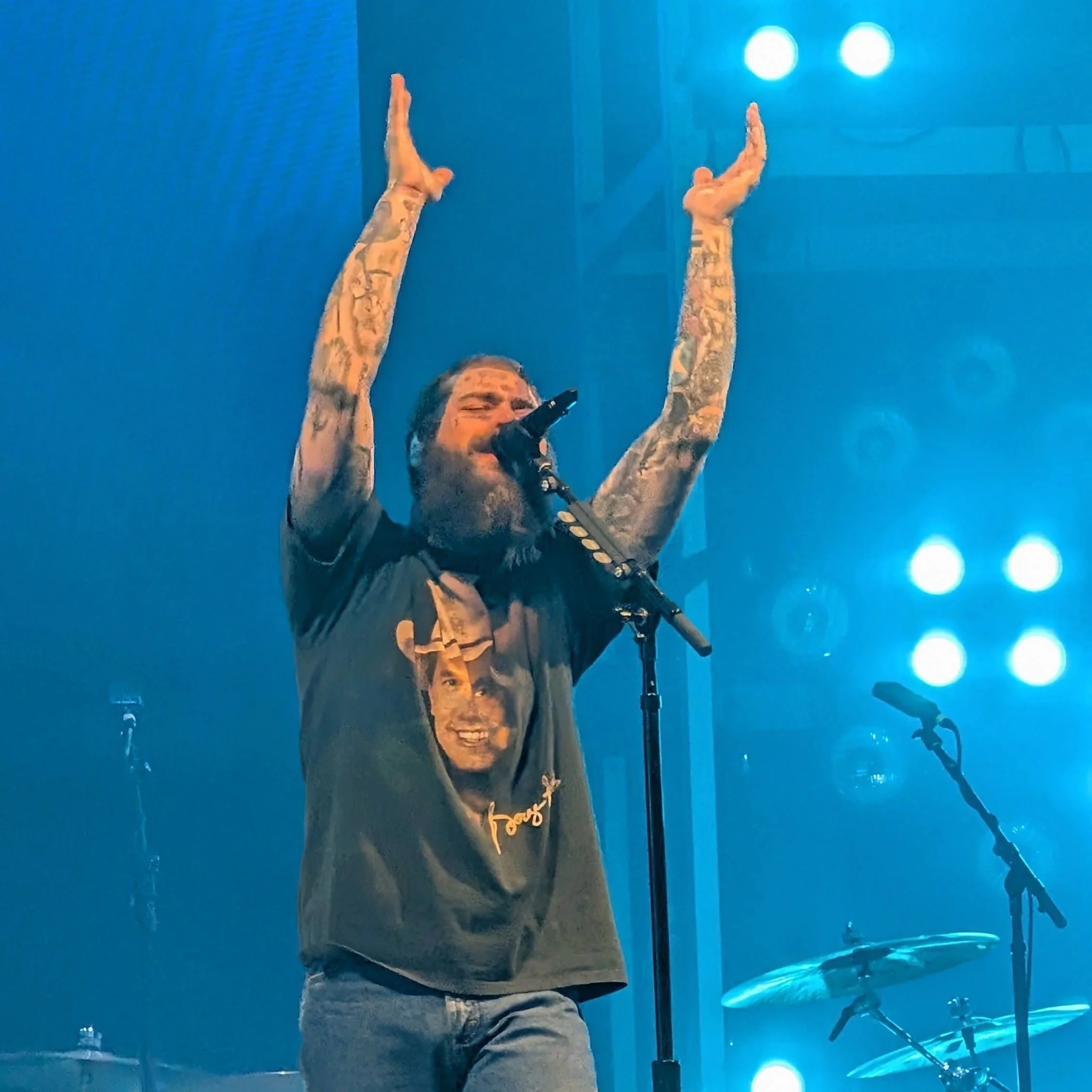 A male singer with tattoos on his arms performing on stage, raising both arms with hands open, singing into a microphone, with blue stage lights behind him.