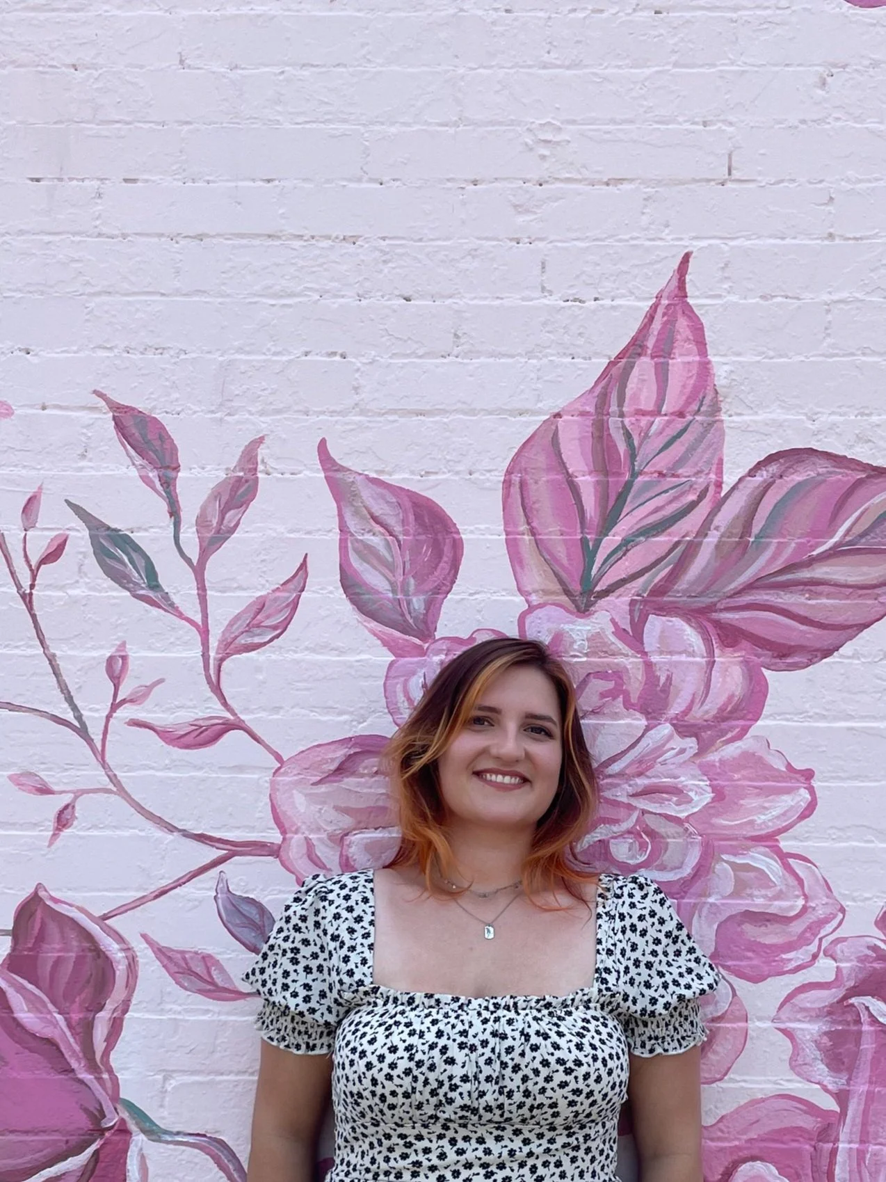 A woman with shoulder-length wavy hair smiling, standing in front of a pink floral mural painted on a brick wall.