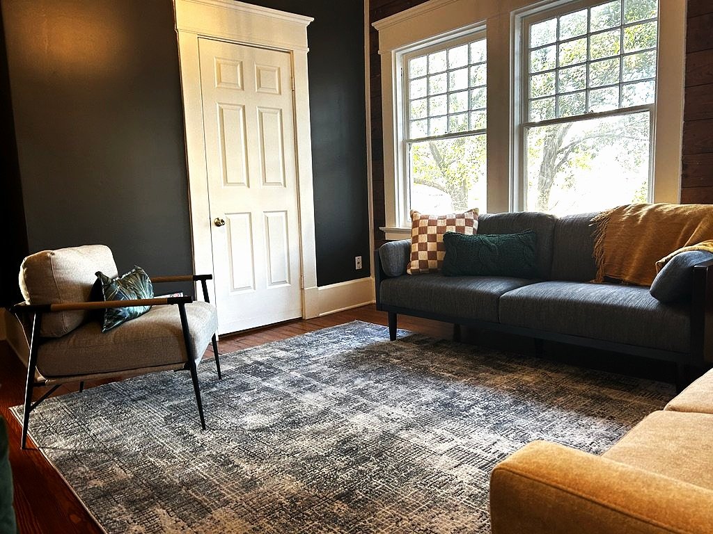 Cozy living room with a dark gray sofa, a beige armchair, and a patterned area rug. Large windows show trees outside. There are decorative pillows and blankets on the furniture, a white door, and wood-paneled wall.