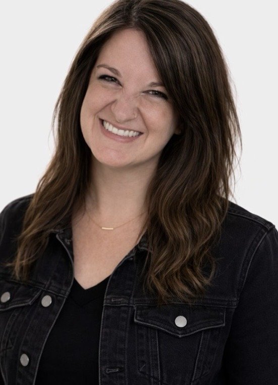 A smiling woman with brown hair and a black denim jacket, standing against a plain white background.