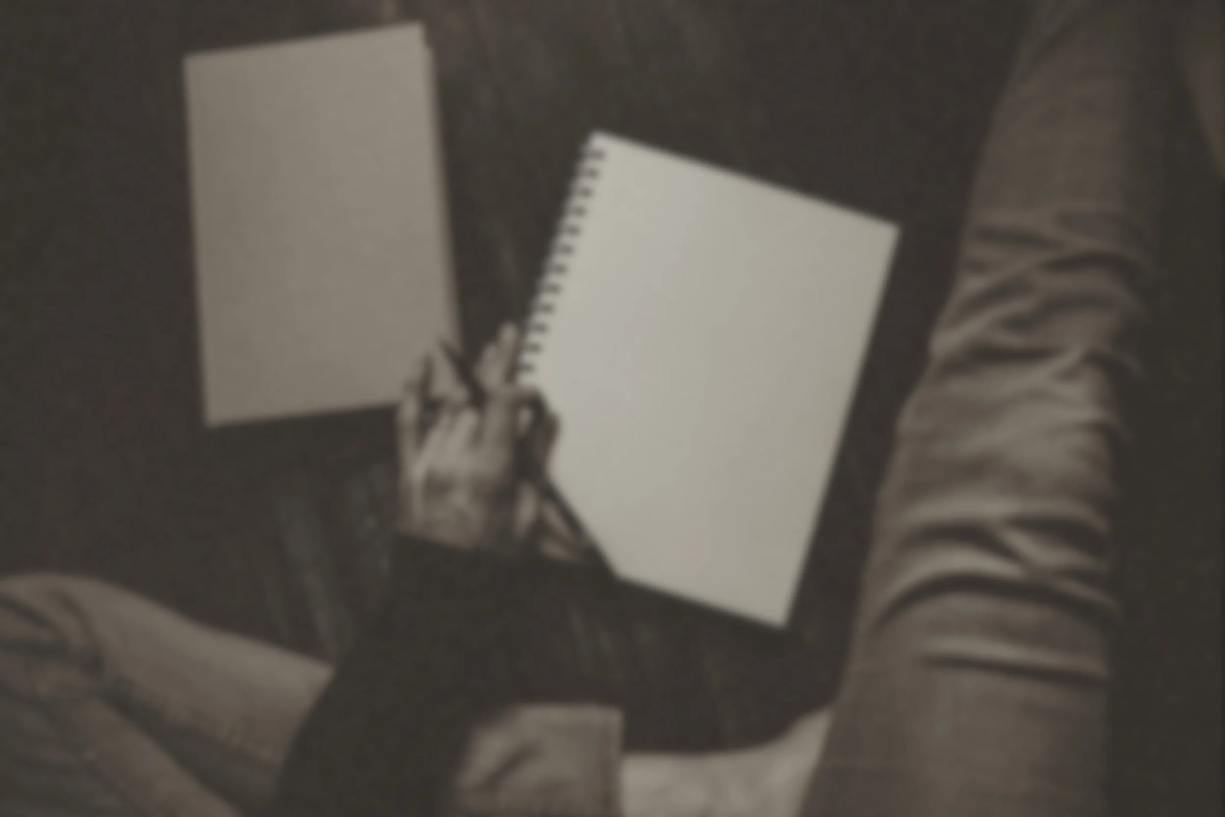 Blurred figure holding blank sketchbook while seated on dark wood floor—an invitation to begin, unfinished and intimate.