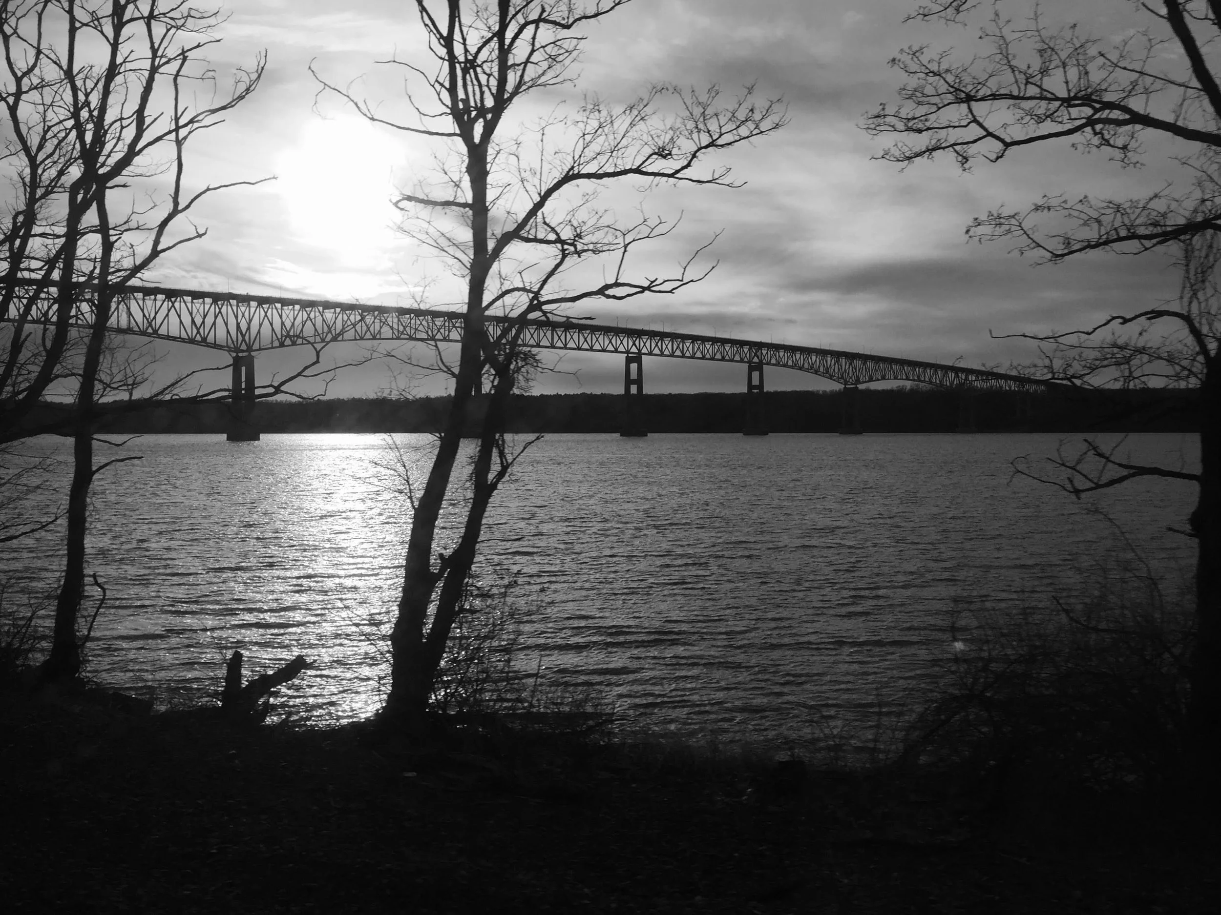 Black and white photo of a bridge spanning across a body of water, with bare trees in the foreground and the sun shining through clouds.