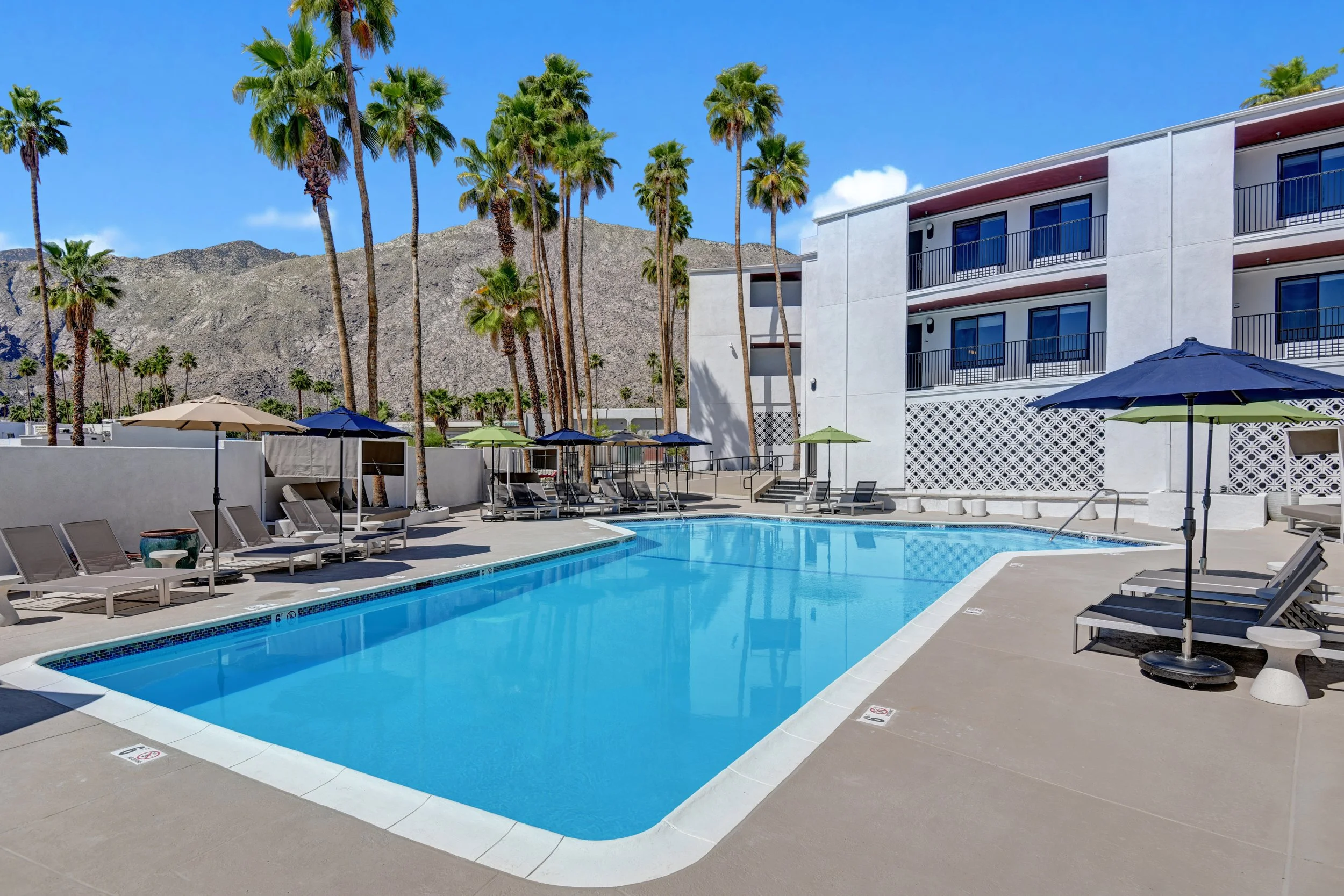 Vacation resort with a swimming pool, palm trees, lounge chairs, umbrellas, and a white hotel building with balconies, mountains in the background, and clear blue sky.