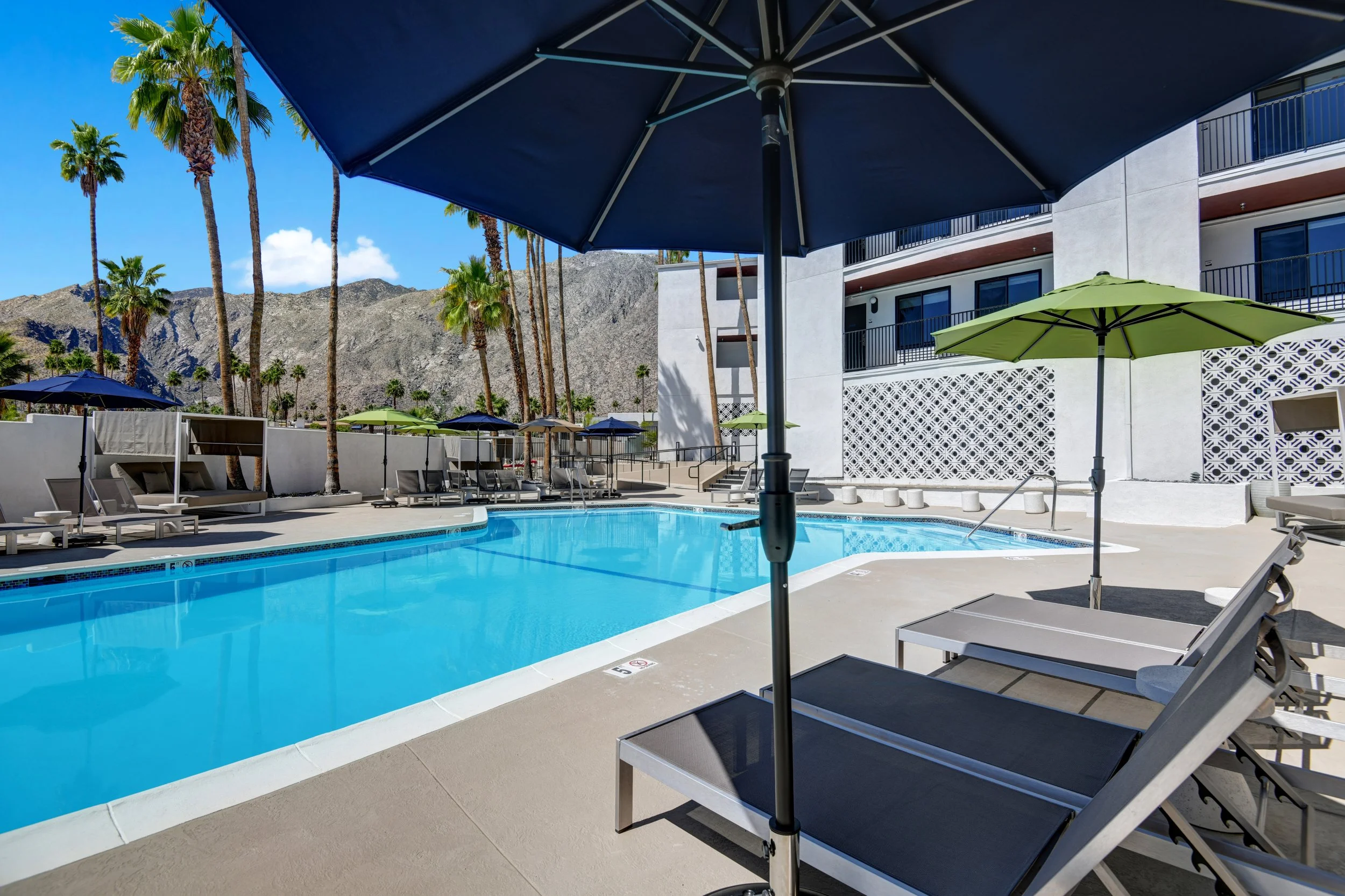 Empty swimming pool with lounge chairs and umbrellas in an outdoor hotel area, with mountains and palm trees in the background.