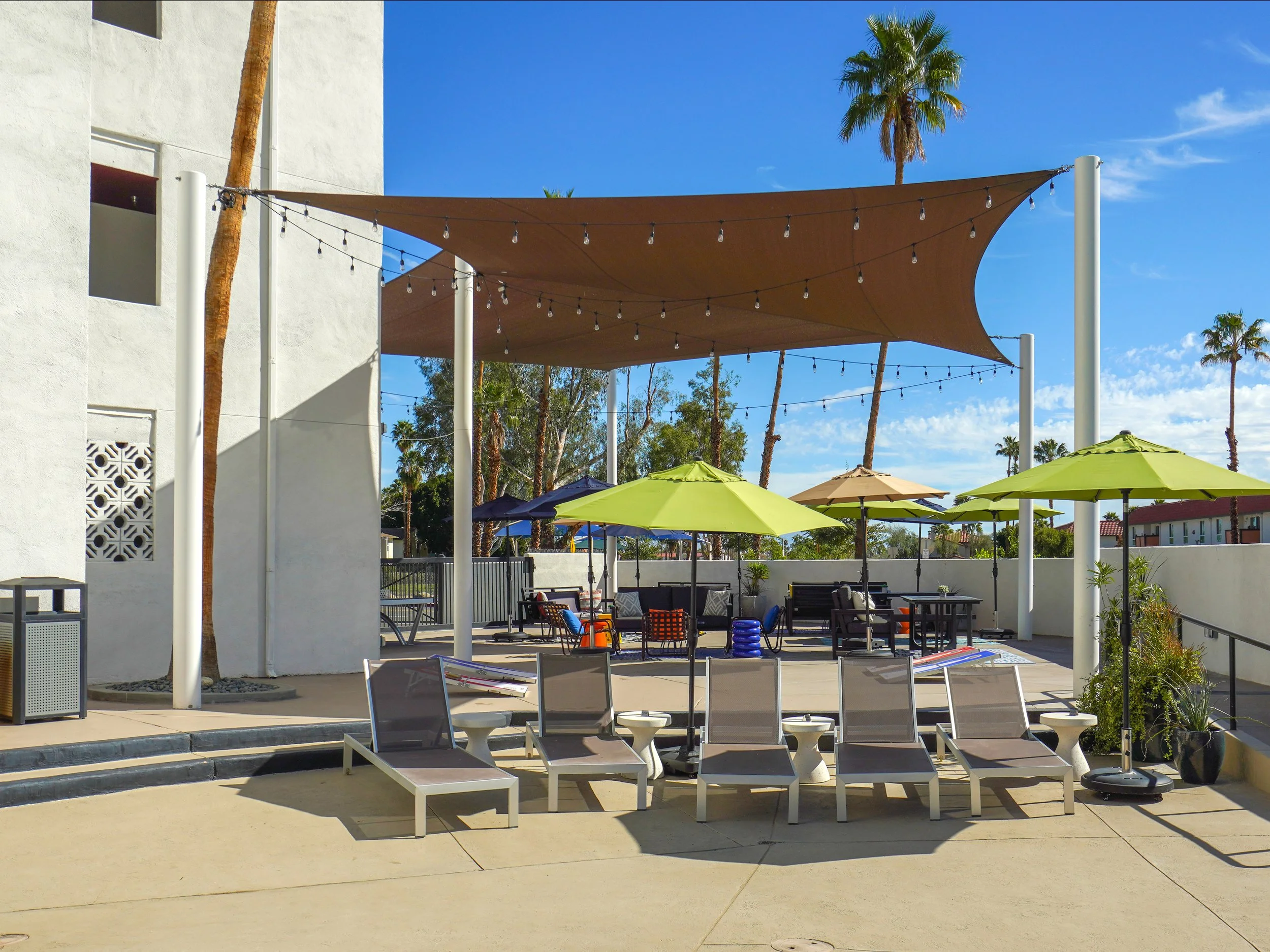 Outdoor poolside patio with lounge chairs, green umbrellas, and a shaded area with string lights and palm trees under a blue sky.