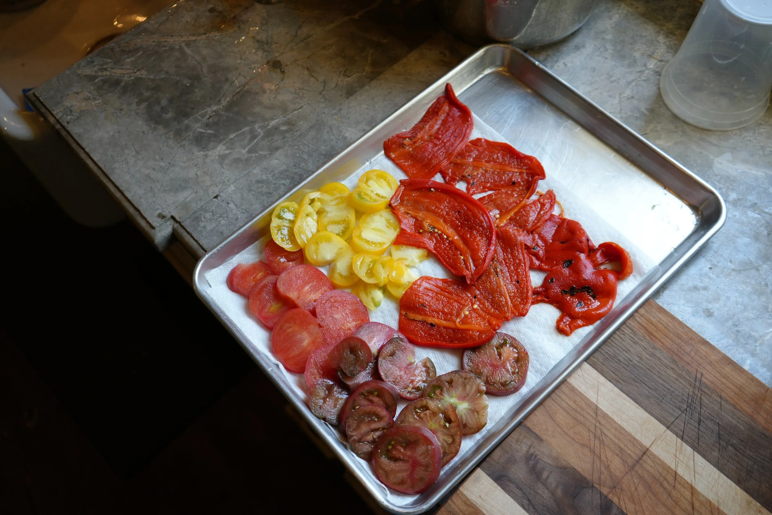 A metal tray holding sliced red and yellow tomatoes on a paper towel, placed on a kitchen counter with a stone surface.