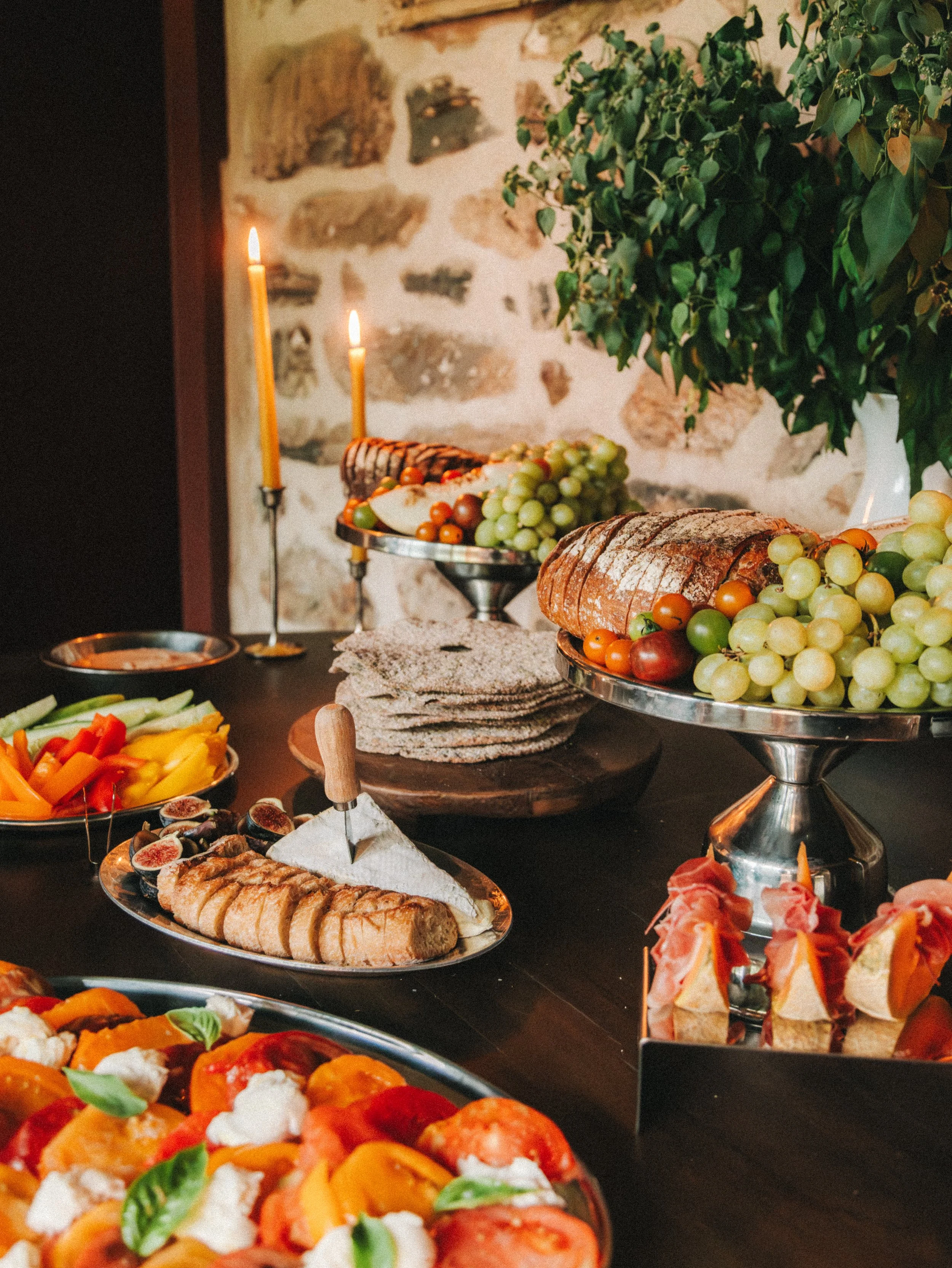 A table set with an assortment of foods including cured meats, cheese, bread, fresh vegetables, and fruit. There are two raised dessert stands with grapes and sliced loaf bread, and a platter of sliced vegetables. Two lit candles are also on the tabl