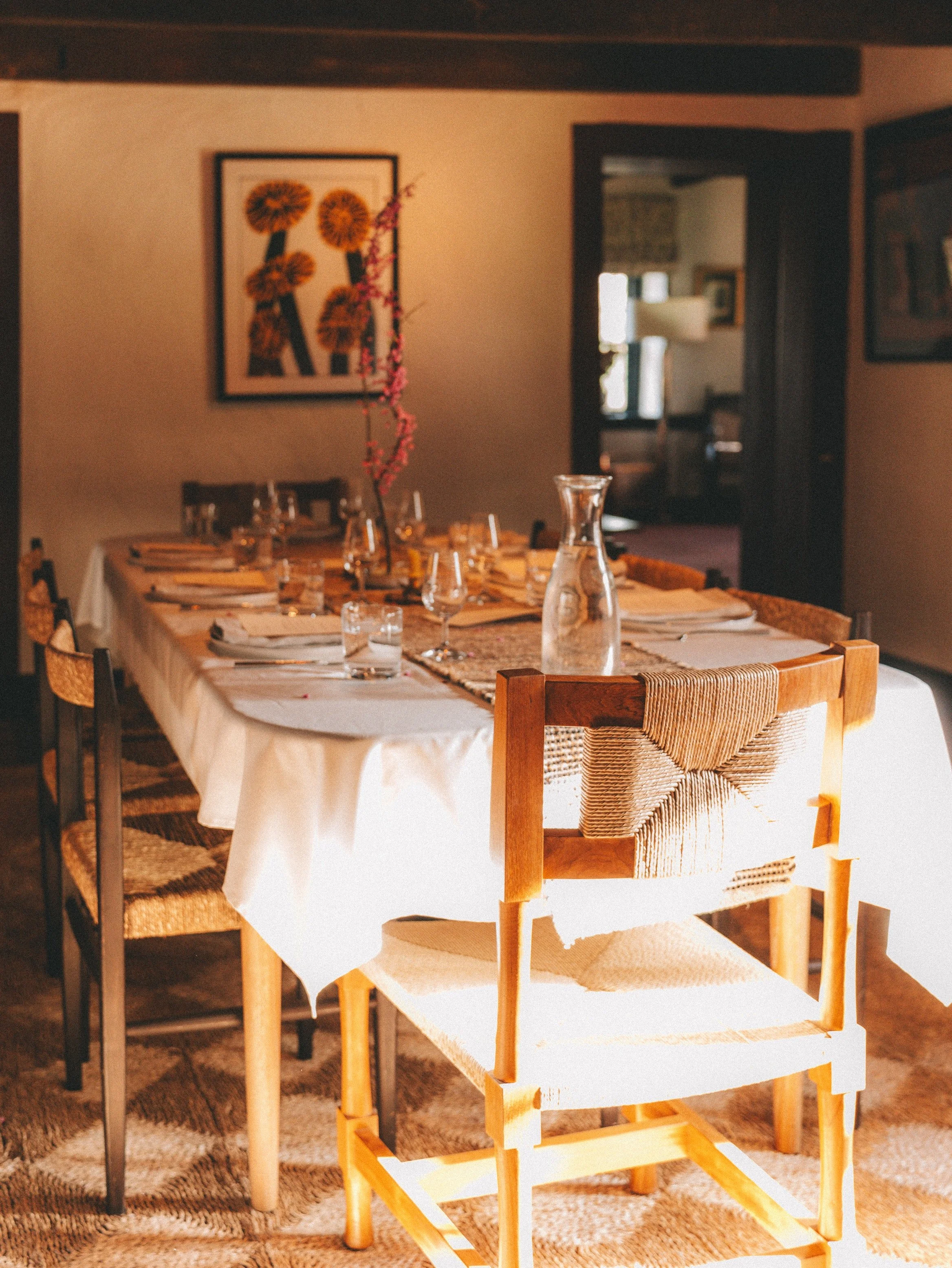 A dining room with a long table set for a meal, decorated with glasses, plates, and a tall pink flower in a branch. There are chairs around the table and framed artwork on the wall.