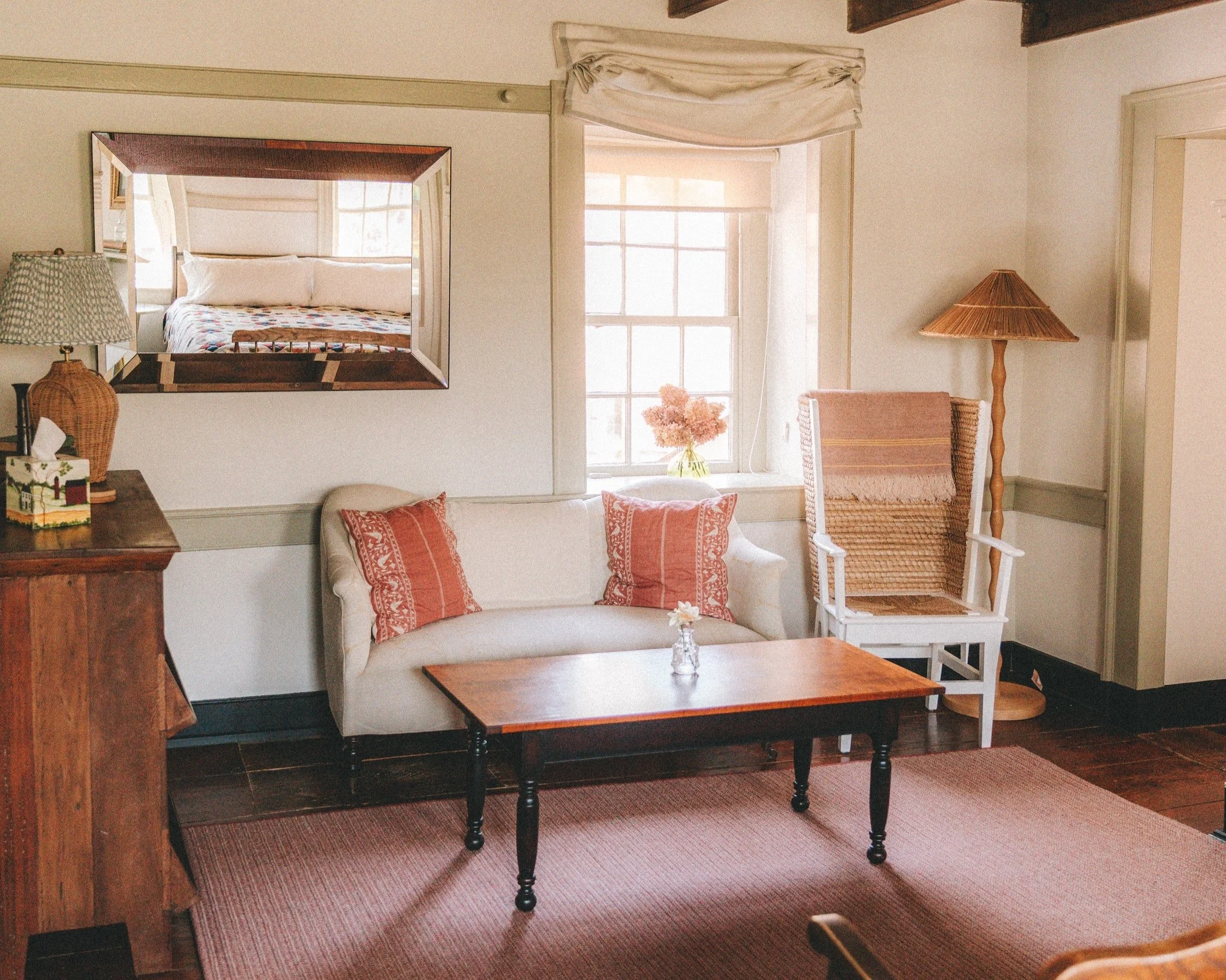 Cozy living room with a white couch, pink pillows, a wooden coffee table, wicker armchair with a pink pillow, and a floor lamp with a wicker shade. There is a window with sheer curtains, a vase of pink flowers on the windowsill, and a large mirror re