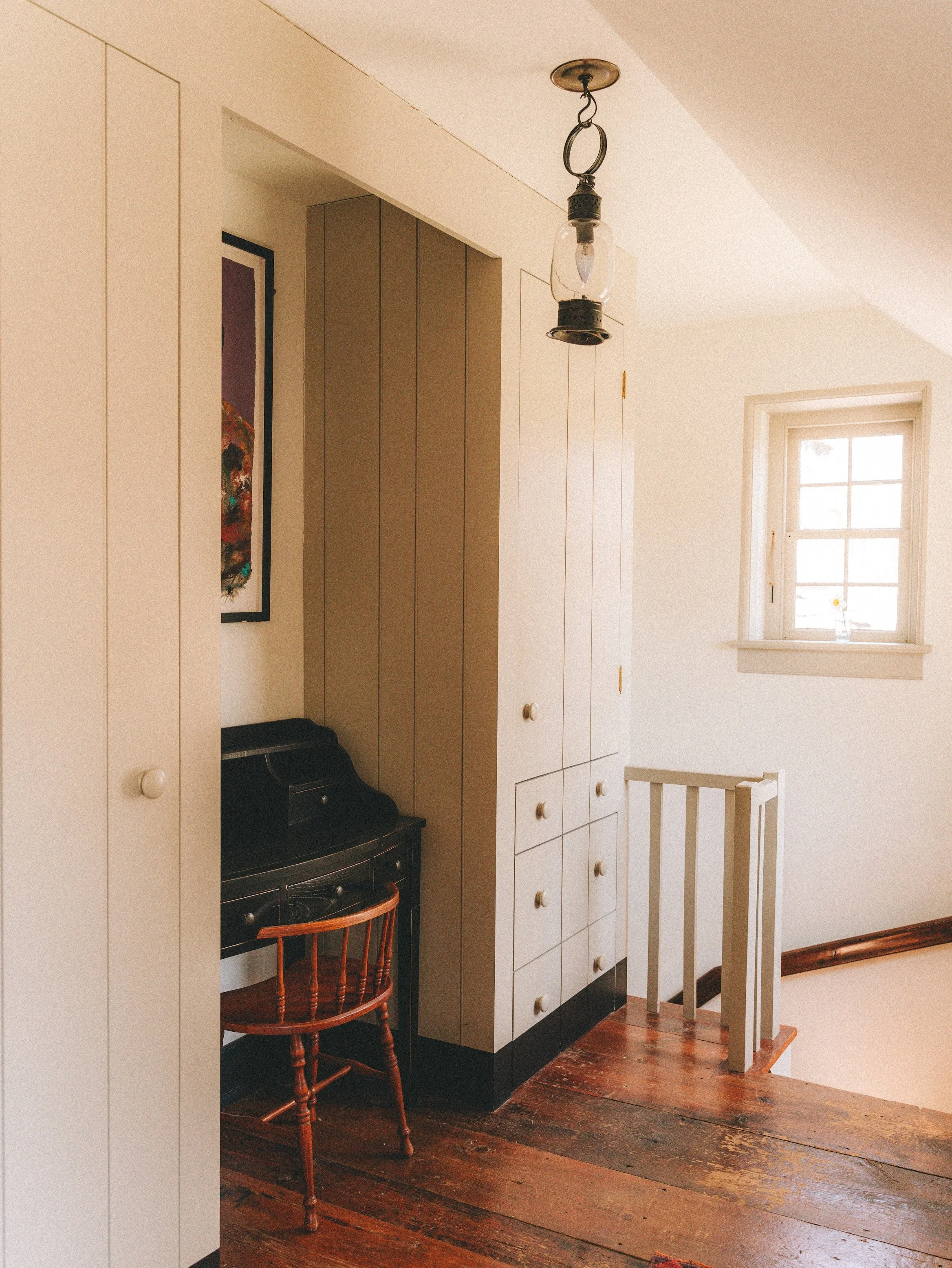 Interior of a house with wooden floor, a small black desk and chair, white cabinets, a window with a flower vase, and a hanging light fixture.