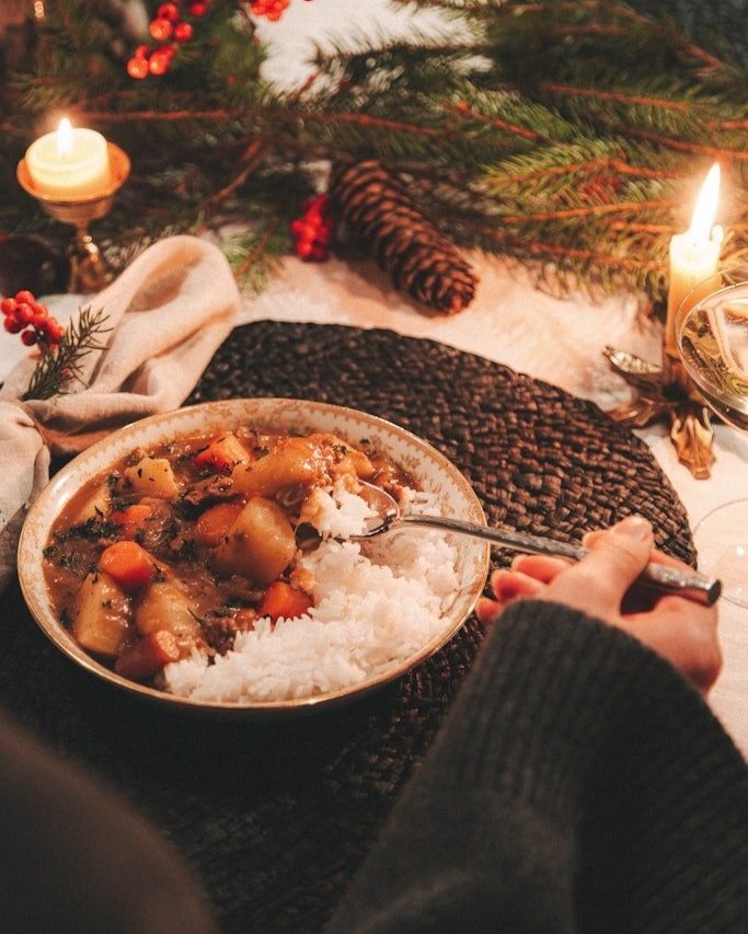 A person serving a bowl of stew with vegetables and rice on a decorated holiday table with pine branches, candles, and a pine cone.