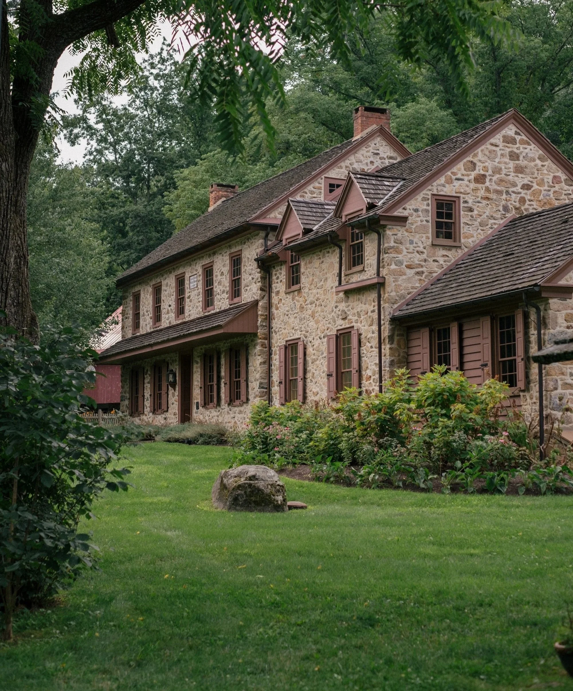 A large stone house with multiple chimneys, surrounded by lush green trees and a well-maintained lawn.