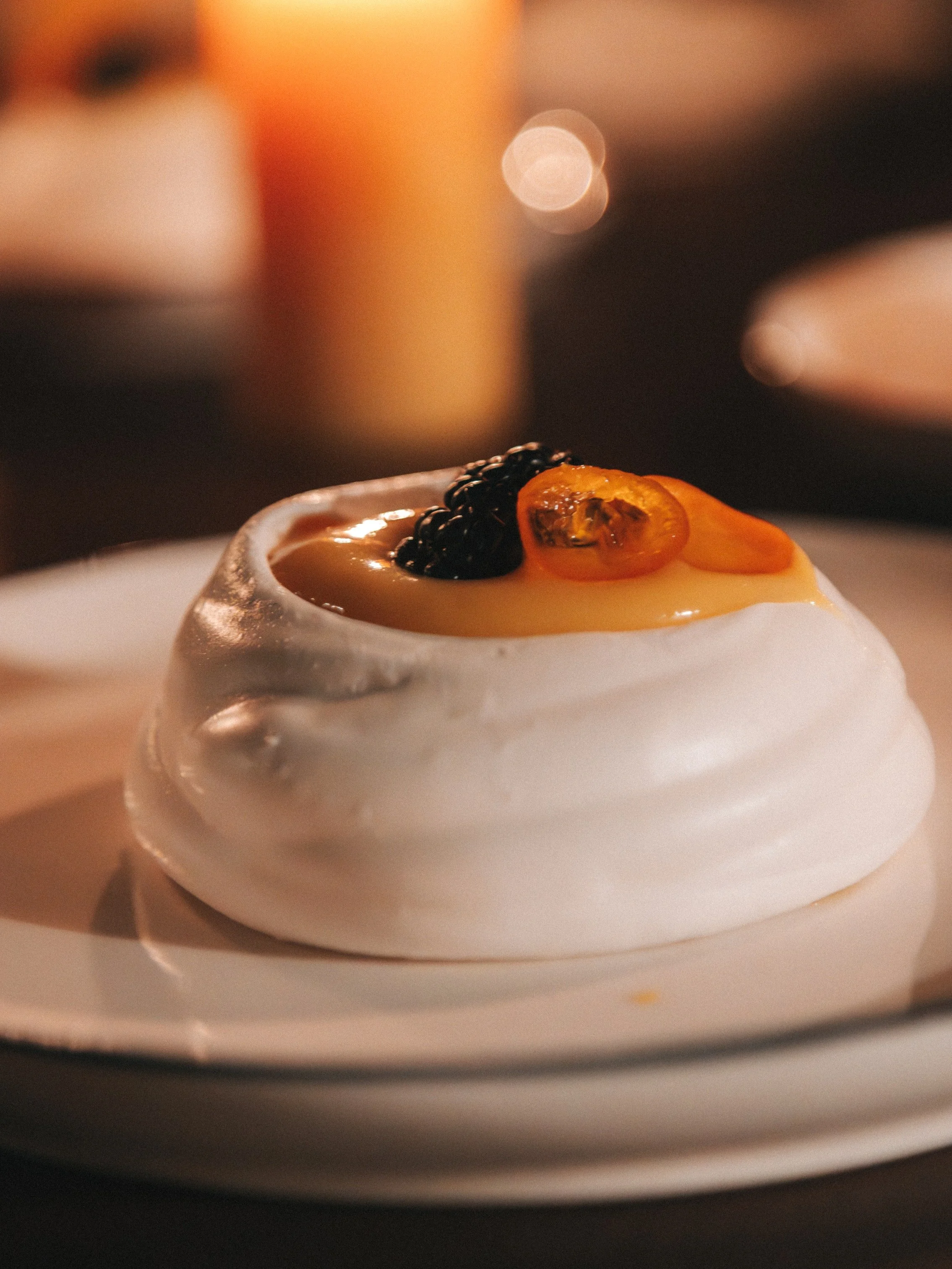 A close-up of a pavlova dessert topped with whipped cream, blackberries, and sliced kumquats on a white plate.