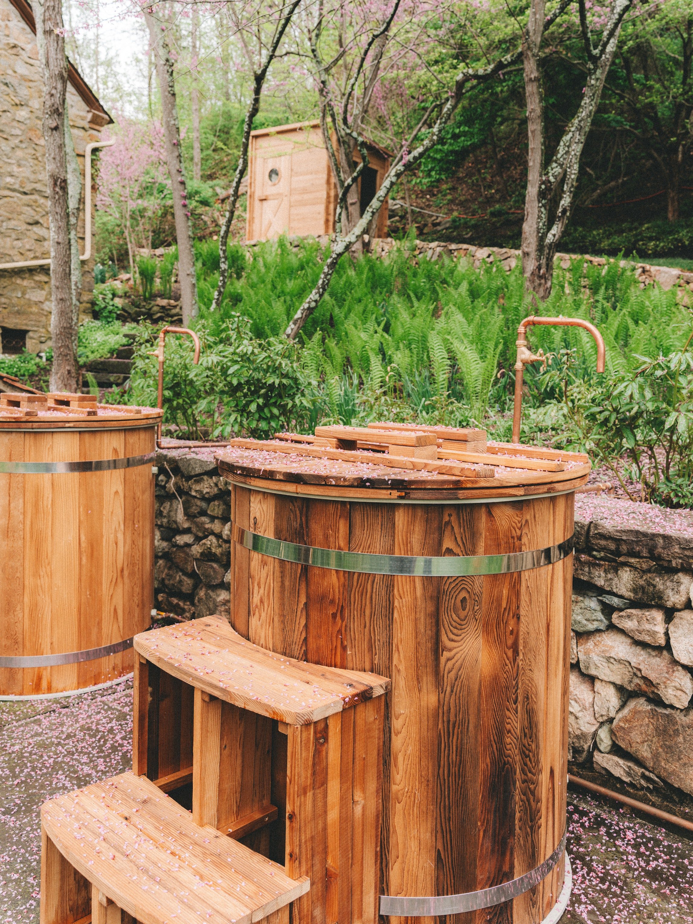 Two wooden hot tubs with copper faucets in a garden with pink flowering trees and a stone wall in the background.