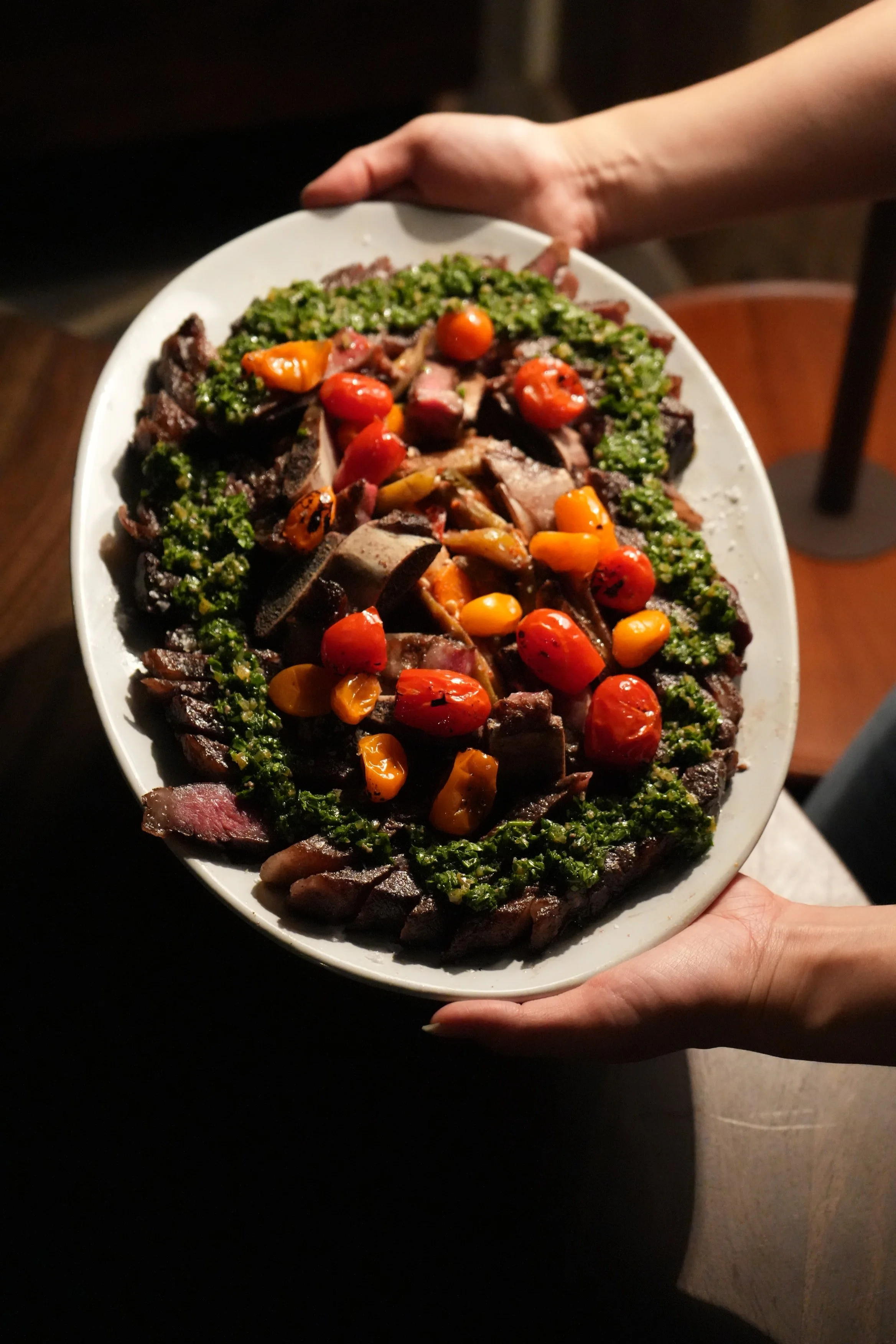 A large oval white platter of sliced steak topped with roasted cherry tomatoes and garnished with chopped herbs.