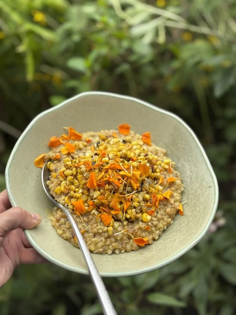 A bowl of cooked cornmeal porridge topped with orange edible flower petals, held outdoors against green foliage.