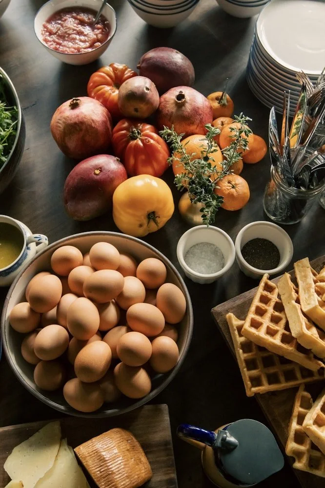 A breakfast table with eggs, pomegranate, tomatoes, a yellow squash, cheese, waffles, syrup, and various condiments.