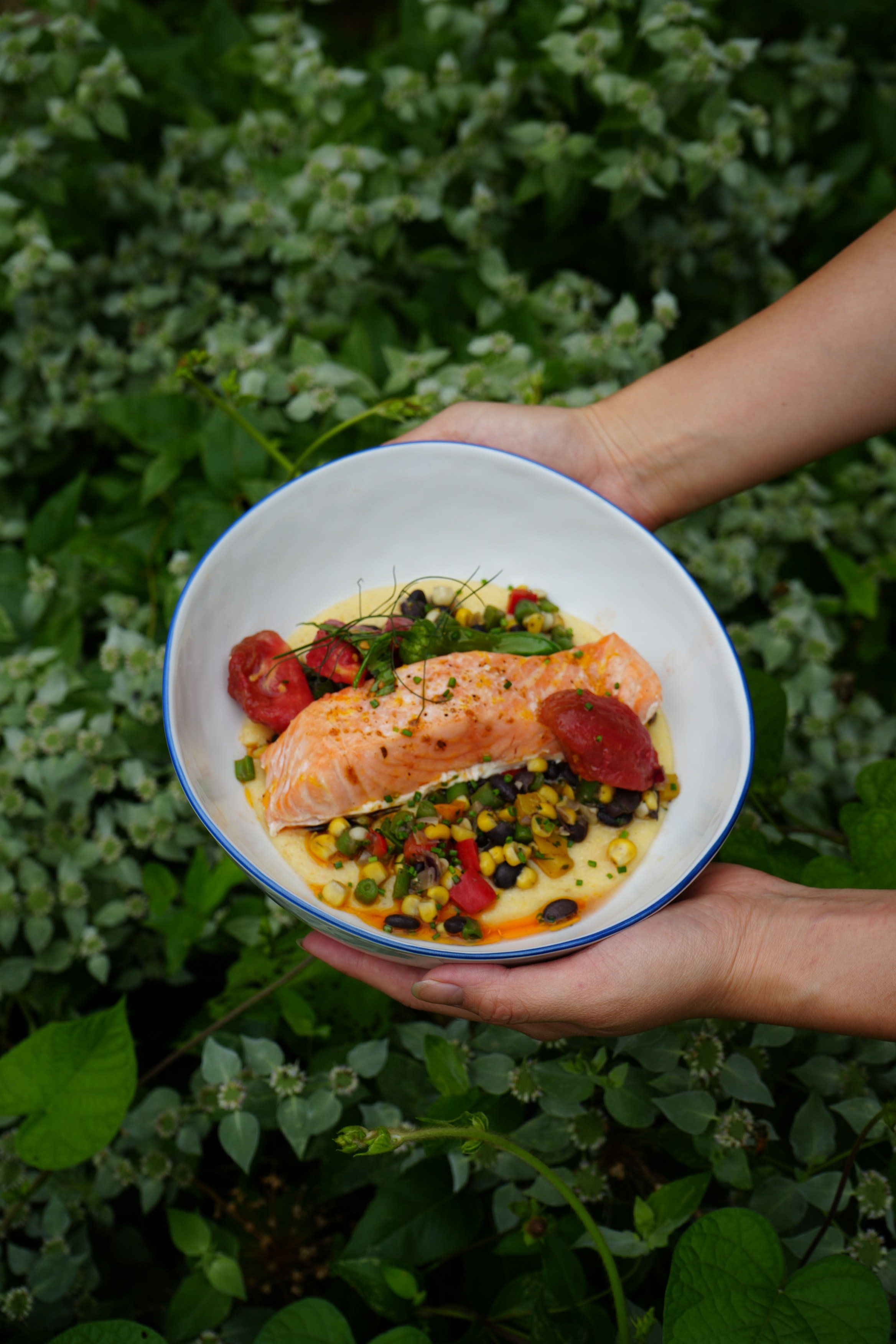 A person holding a white bowl with a salmon fillet atop a bed of mixed vegetables and corn, garnished with herbs, against a background of green foliage.