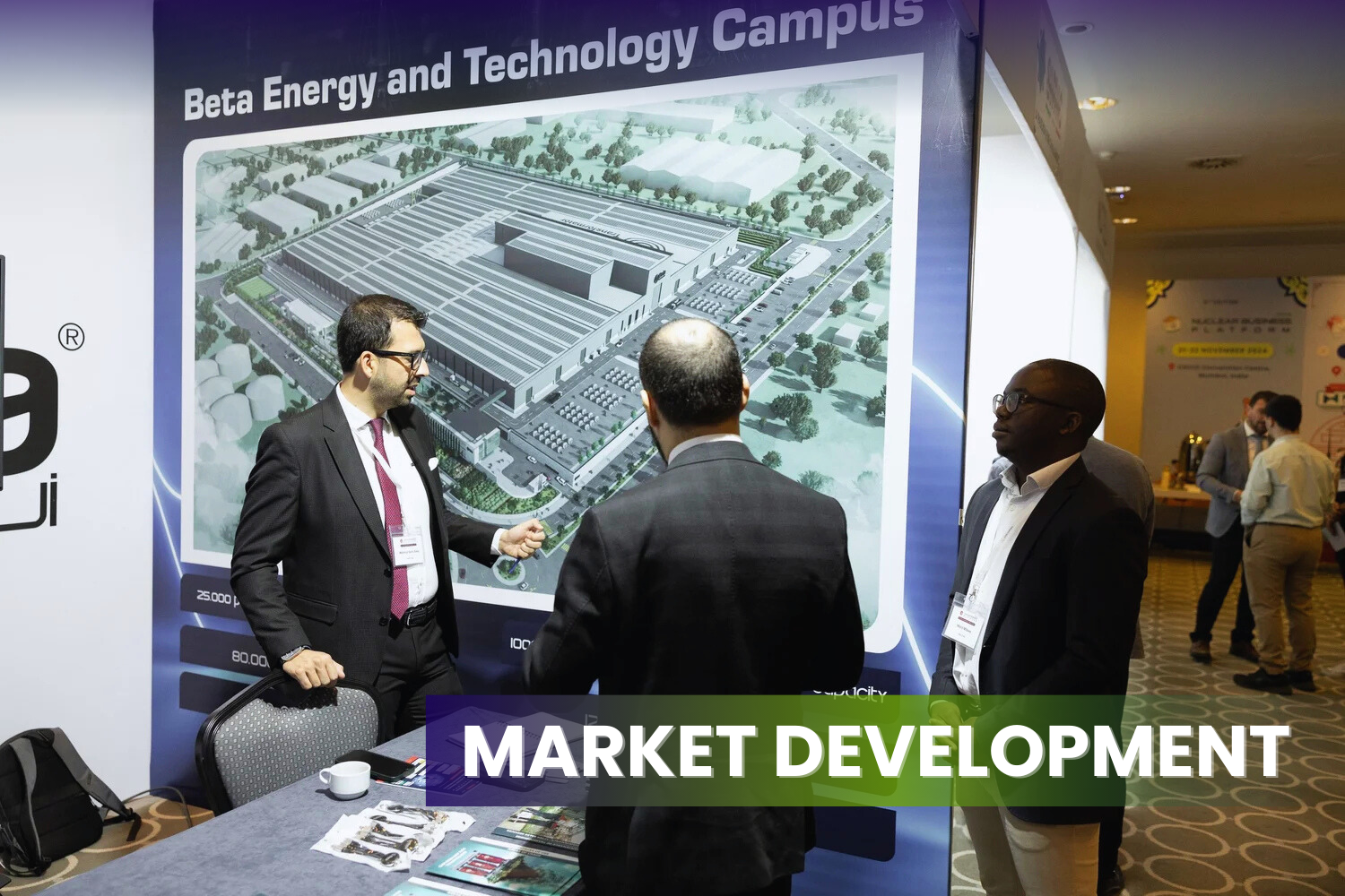 Three men in suits having a discussion at a booth with a large display showing the layout of a 'Beta Energy and Technology Campus' at a business conference or exhibition.