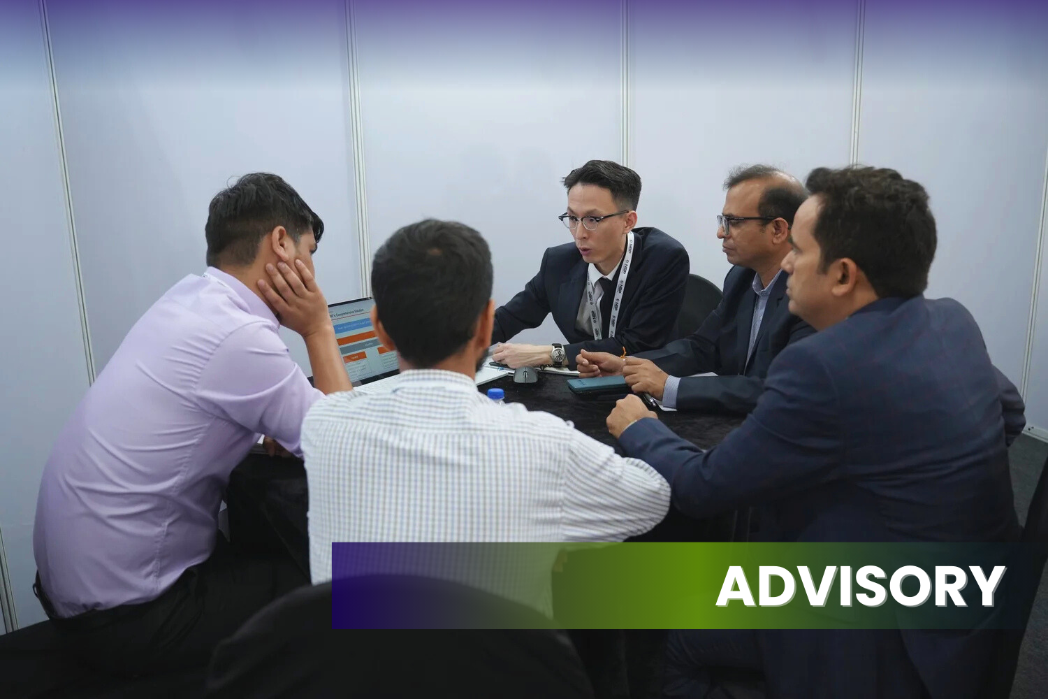 Five men having a serious meeting in a small conference room, with one person explaining something to the group, and a laptop open on the table. The word 'ADVISORY' is displayed at the bottom right of the image.