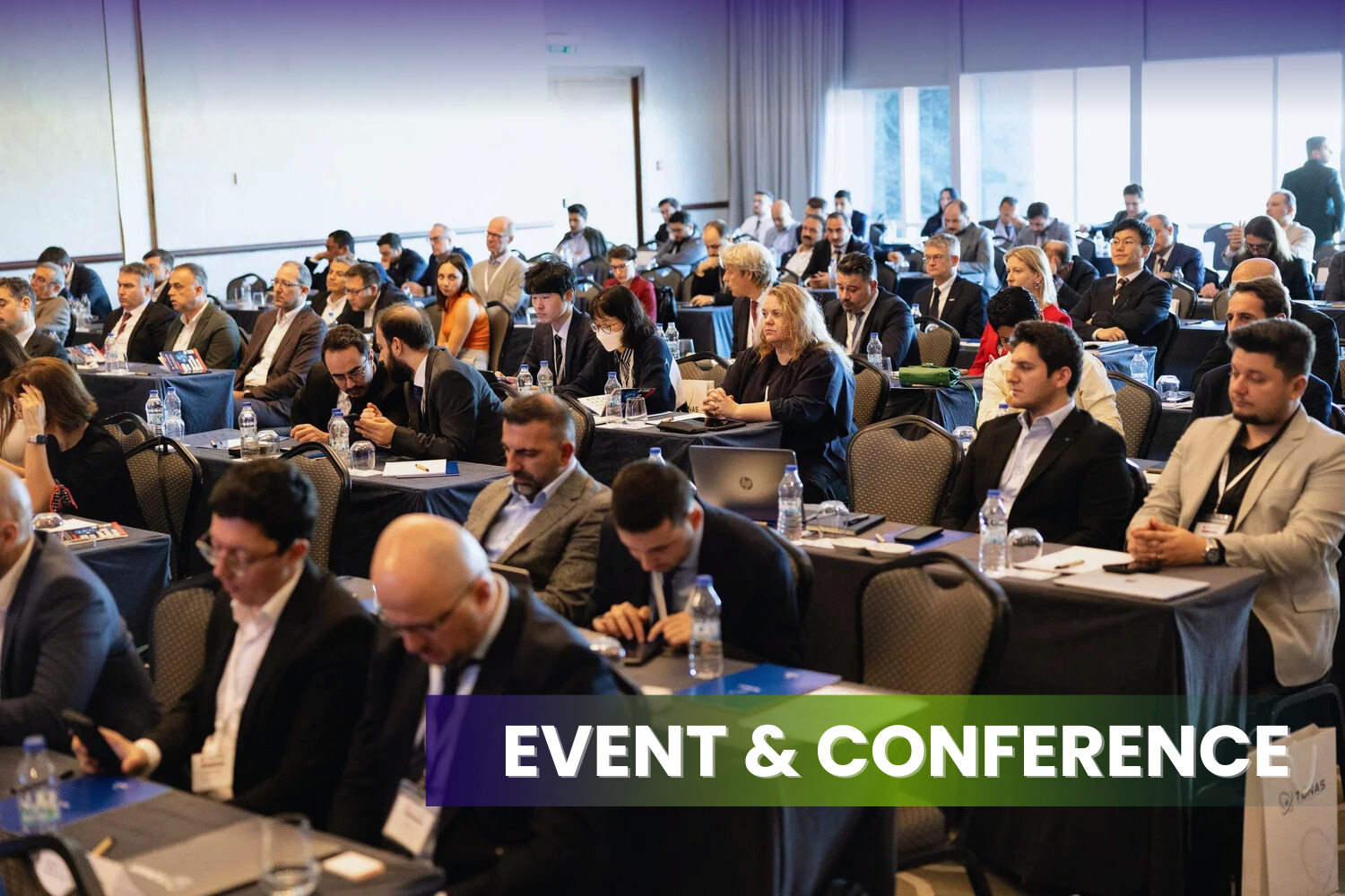 Large group of diverse professionals attending a conference in a spacious, well-lit room with large windows, sitting at tables with water bottles, notebooks, and laptops, and listening attentively to a speaker.