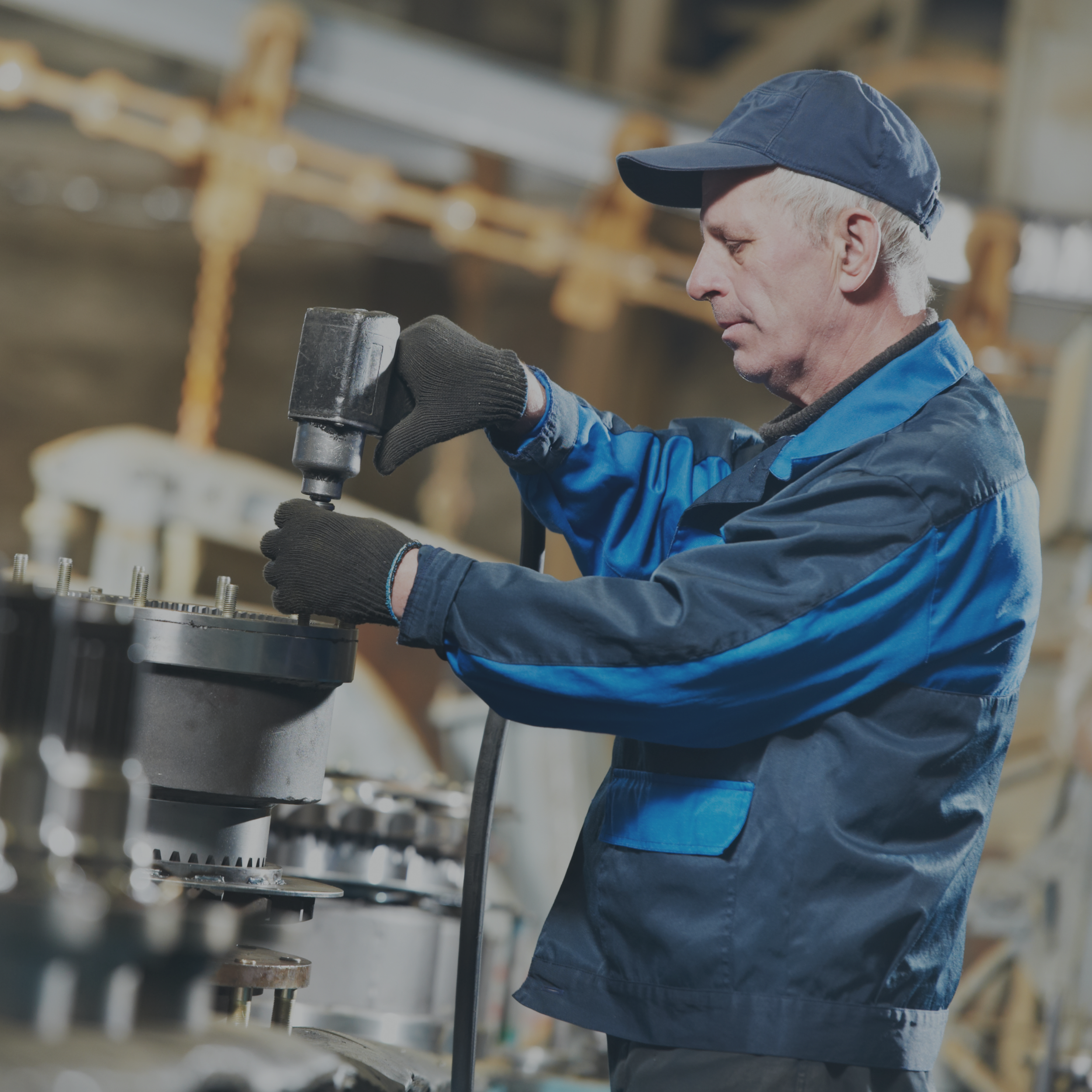 A man wearing a blue cap and blue jacket working with a power tool in a workshop. He appears focused on a piece of equipment or machinery.